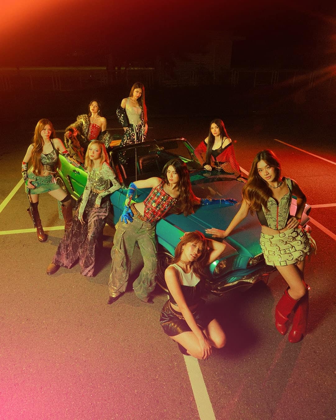 Eight women pose confidently around and on a vintage turquoise car in an empty parking lot at night, illuminated by colorful, stylized lighting with a retro vibe. They wear bold, fashionable outfits and striking poses.