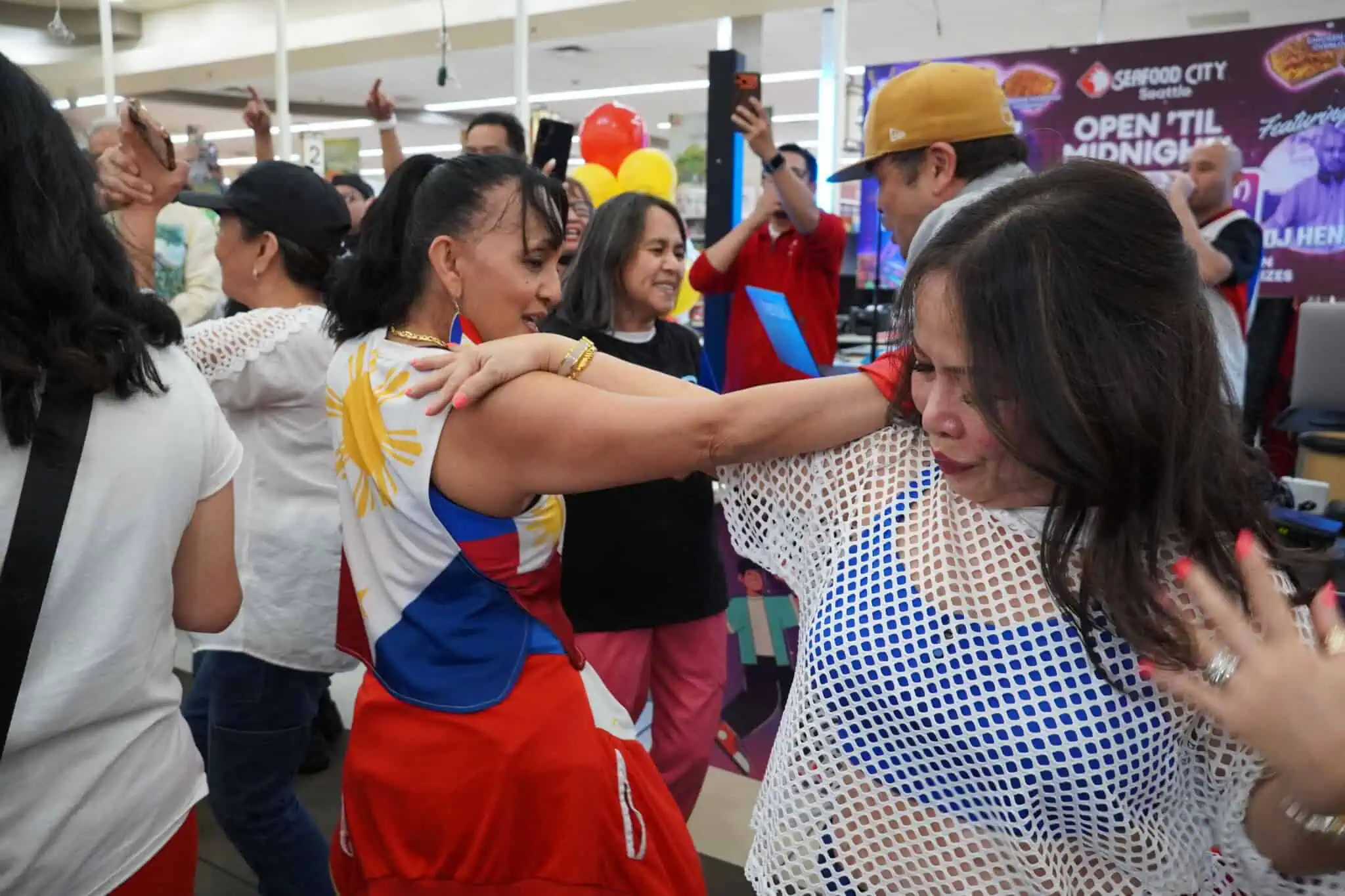 A group of people dance energetically at an indoor event. Two women in front wear colorful outfits and smile, surrounded by others enjoying the lively atmosphere. Balloons and a sign are visible in the background.