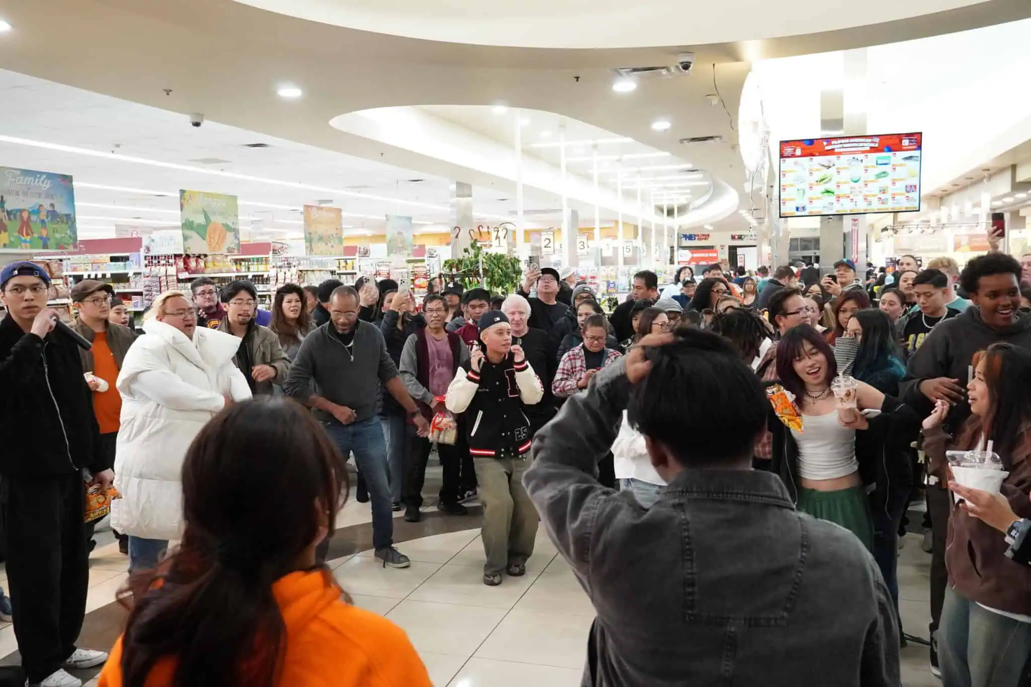 A large, diverse crowd of people gathers indoors, many smiling, laughing, and dancing amidst bright store lighting and colorful signs in the background. The atmosphere appears energetic and festive.