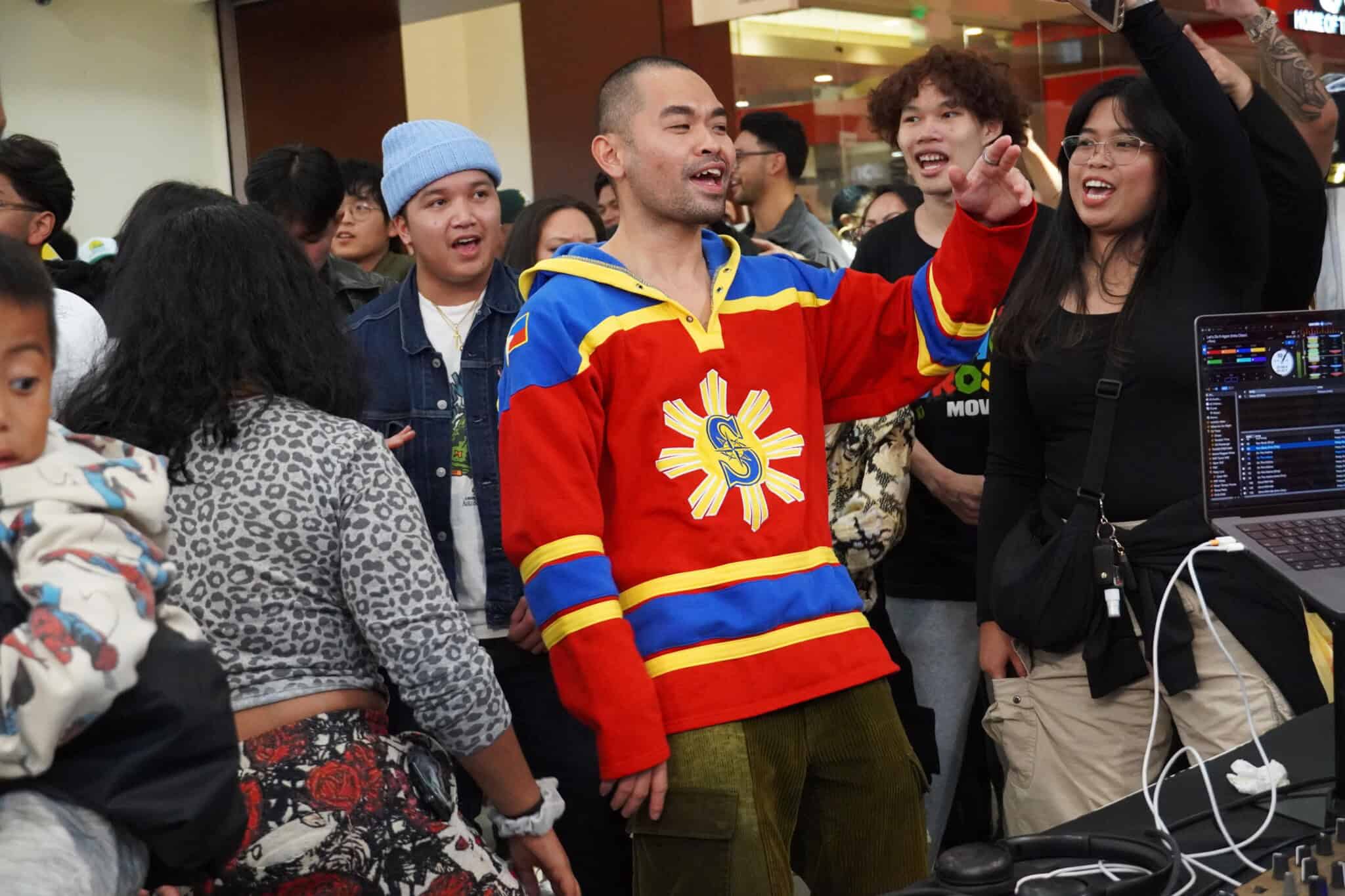 A group of people cheer and smile at an indoor event. One person in front wears a red, yellow, and blue jersey with a sun symbol, raising his arm enthusiastically. A DJ setup is visible on the right.