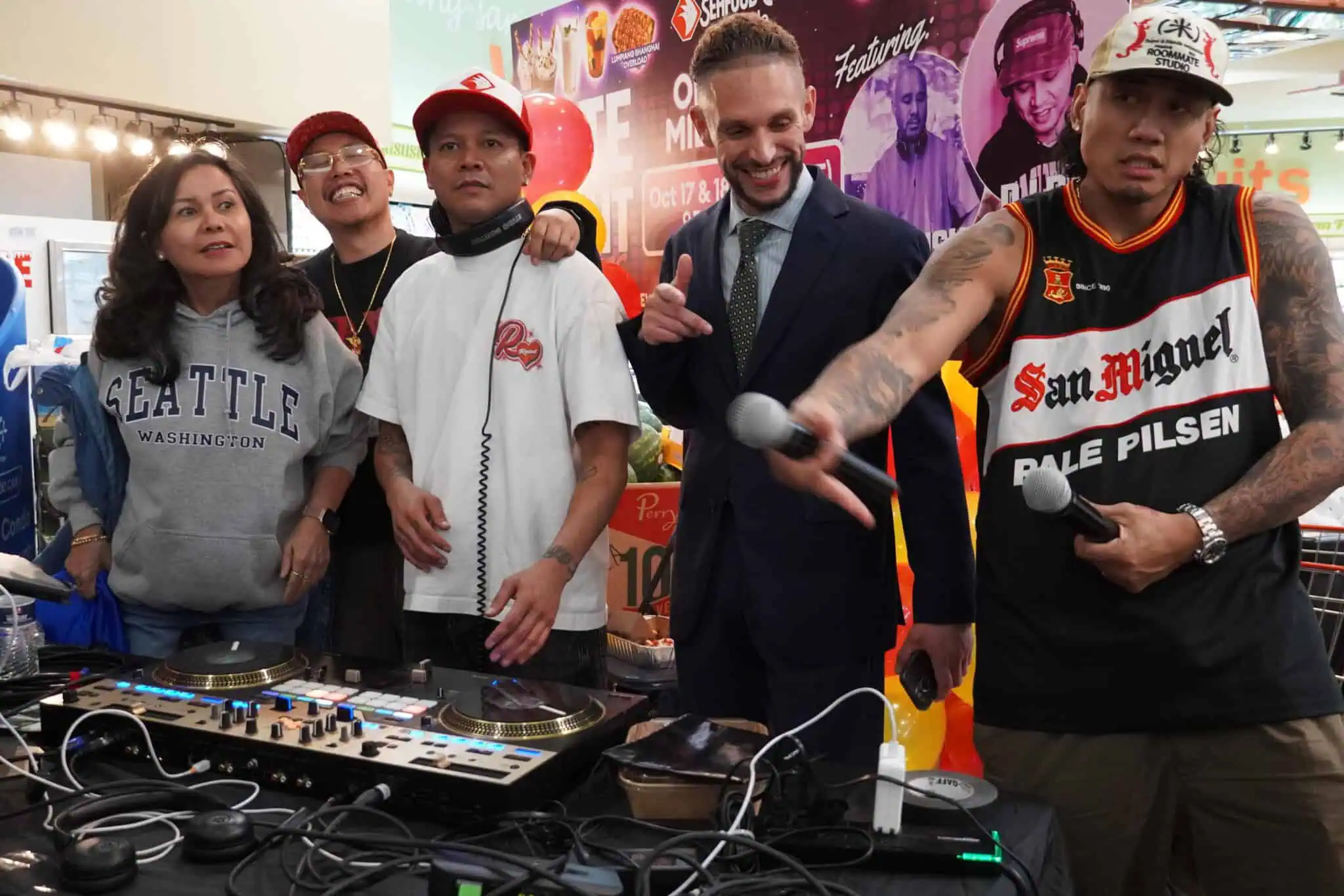 Five people stand together at a DJ booth with equipment. One holds two microphones, another wears headphones and a red cap, and another gestures a peace sign. A colorful, busy background includes posters and lights.