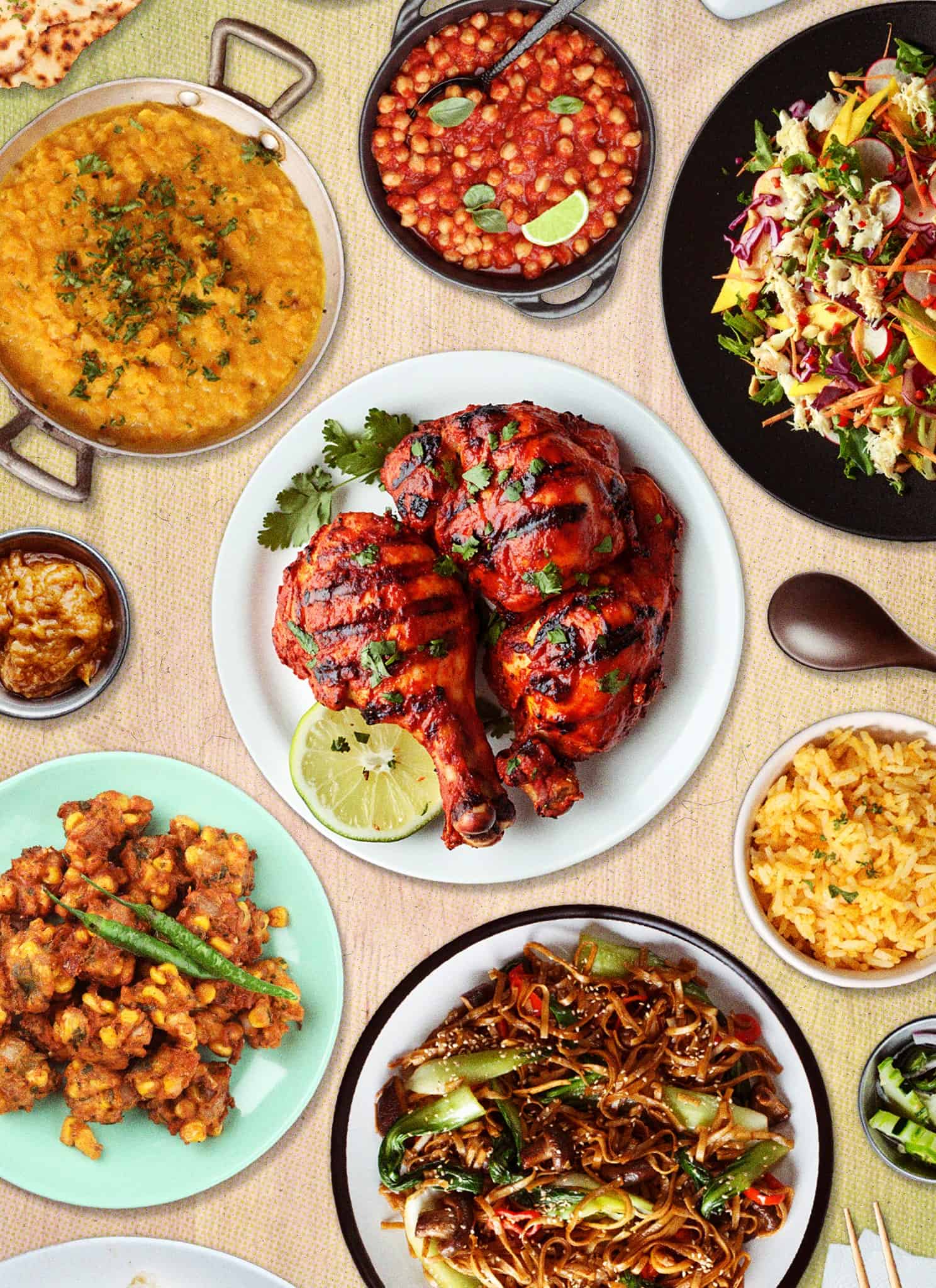 A colorful spread of various Indian dishes, including grilled tandoori chicken, rice, dal, naan, chickpea curry, noodles, stir-fried vegetables, salad, chutney, and fried appetizers, all arranged on a table.
