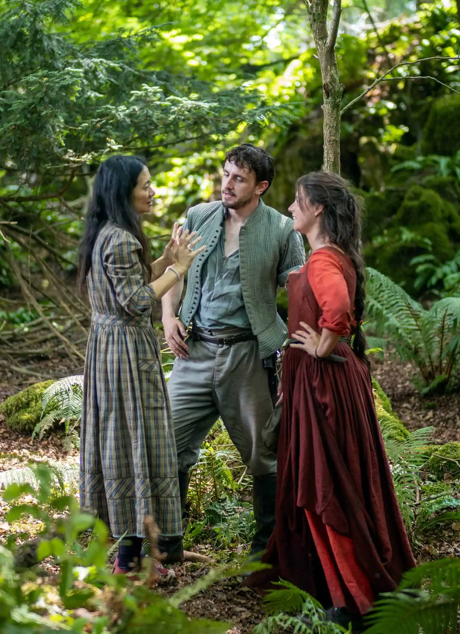 Three people in old-fashioned clothing stand in a lush forest, engaged in conversation. Ferns and moss cover the ground, and sunlight filters through the trees above.
