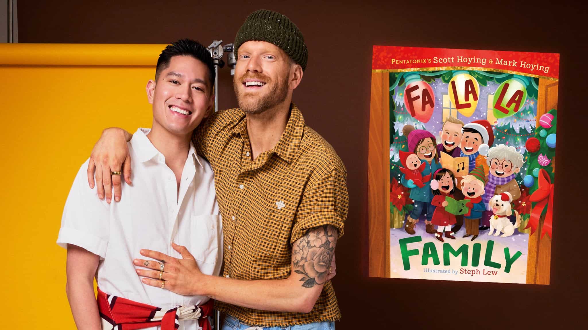 Two smiling men pose together in front of a yellow background. Beside them is the cover of a colorful childrens book titled Fa La La Family, showing a diverse family singing together with festive decorations and pets.