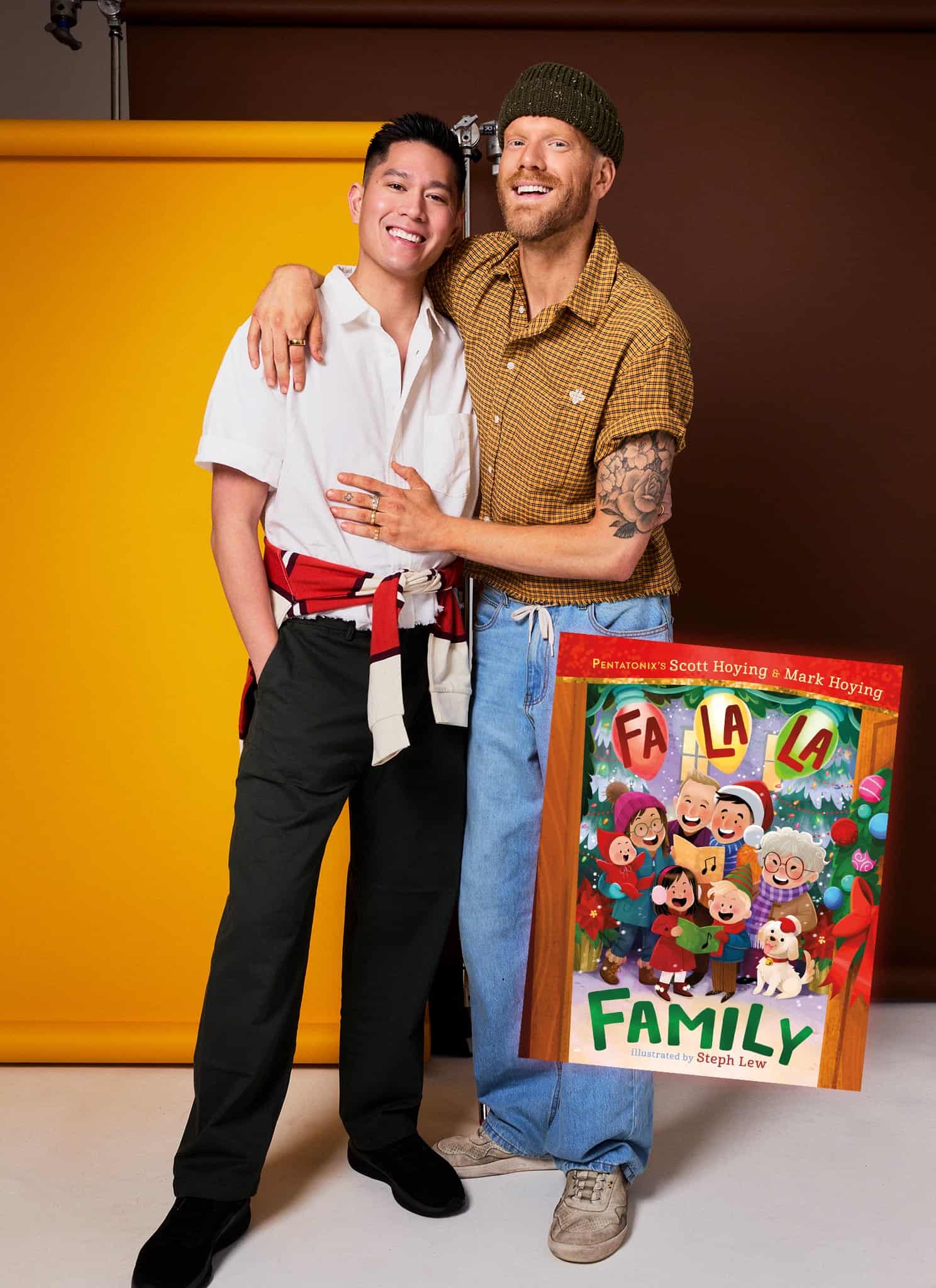 Two smiling men pose together in front of a yellow background. Beside them is the cover of a colorful childrens book titled Fa La La Family, showing a diverse family singing together with festive decorations and pets.