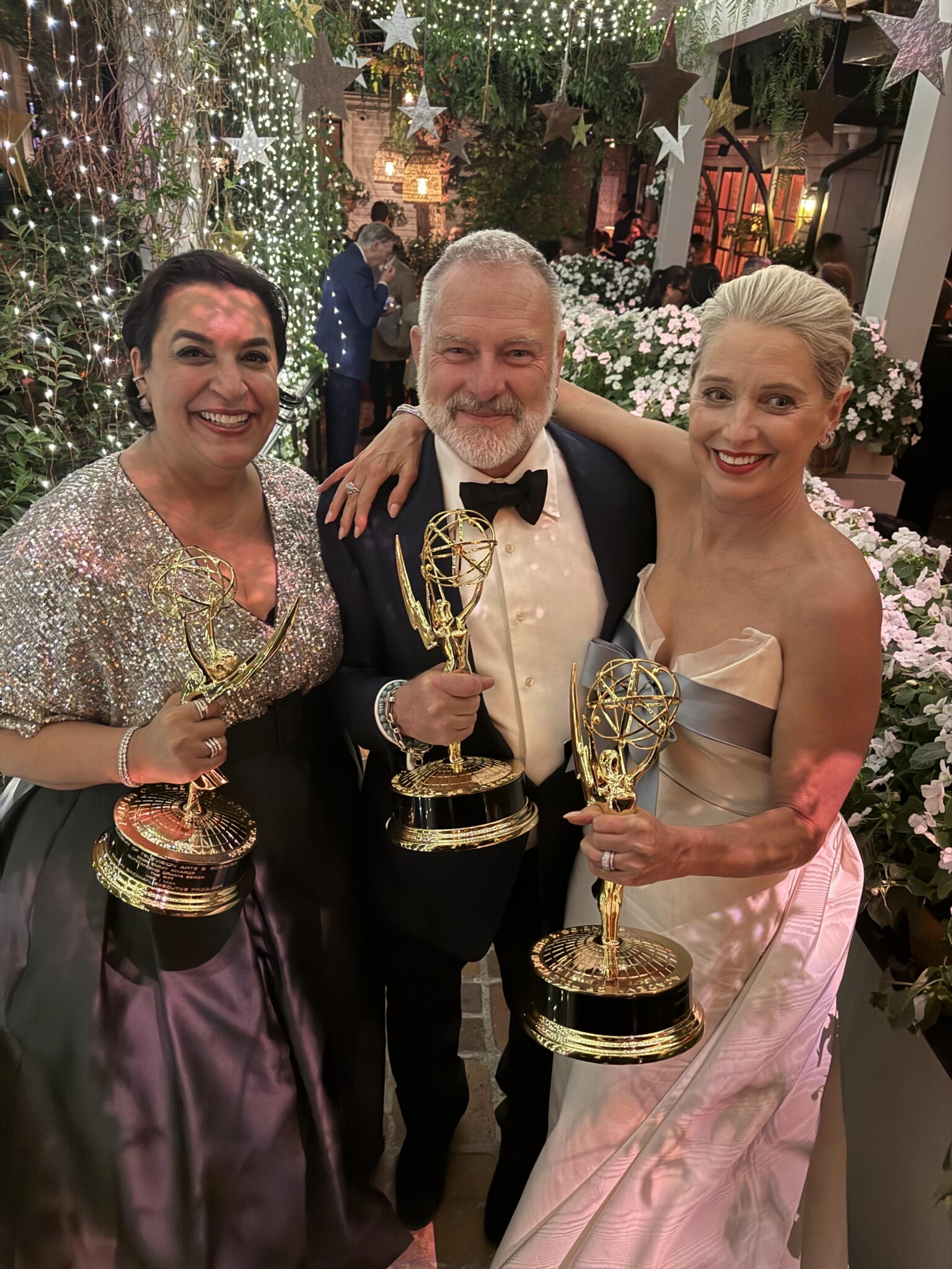Three people dressed in formal attire smile and pose together, each holding an Emmy award, surrounded by flowers and sparkling lights at an outdoor event.