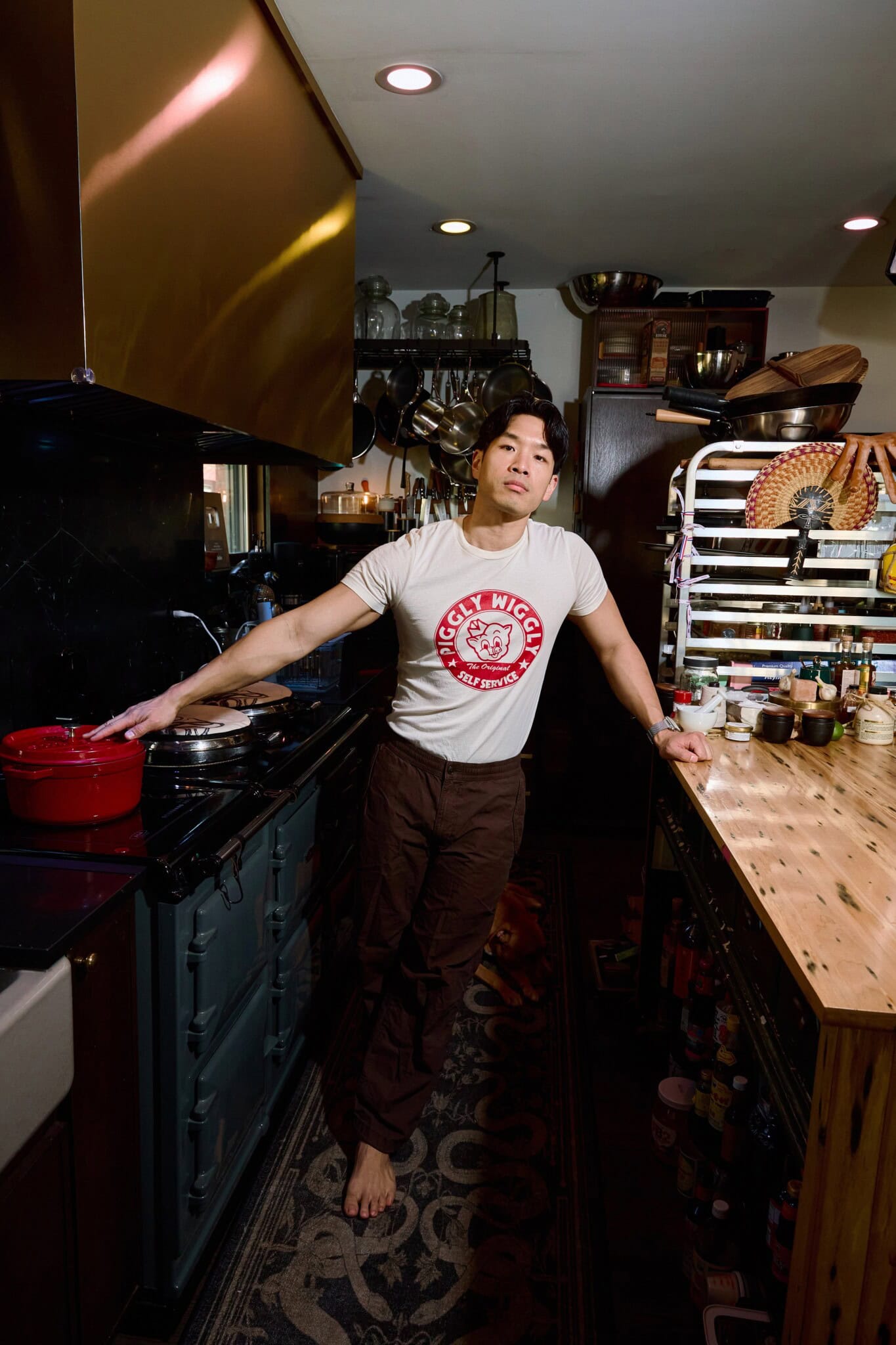 A person in a Piggly Wiggly t-shirt stands barefoot in a cozy, well-stocked kitchen, leaning on a wooden counter with one hand on a red pot on the stove.