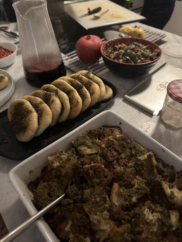 A table with a white surface displaying sliced bread with zaatar, a carafe of red wine, a casserole dish with baked stuffing, a bowl of salad, a pomegranate, and kitchen tools in the background.