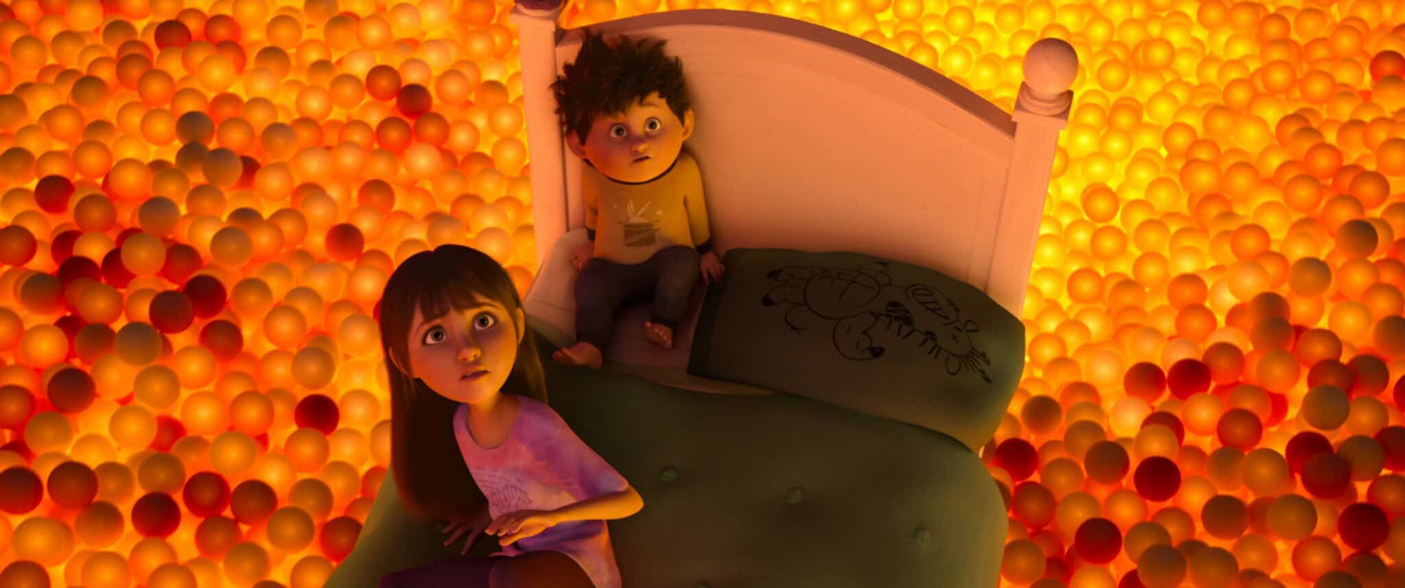 Two animated children sit on a bed surrounded by a sea of glowing orange and yellow balls, looking upward with surprised expressions. The scene is warm and brightly lit.