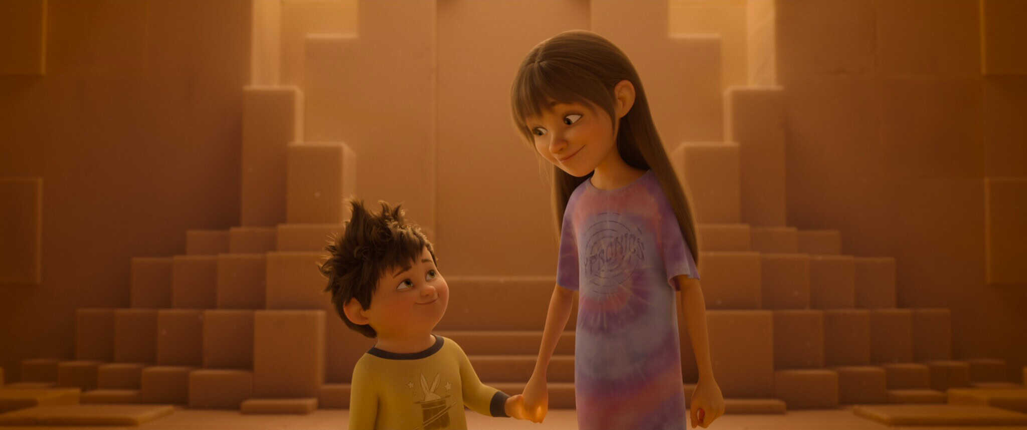 A smiling young boy with messy hair holds hands with an older girl in a tie-dye shirt, both standing in a softly lit, geometric room with steps in the background.