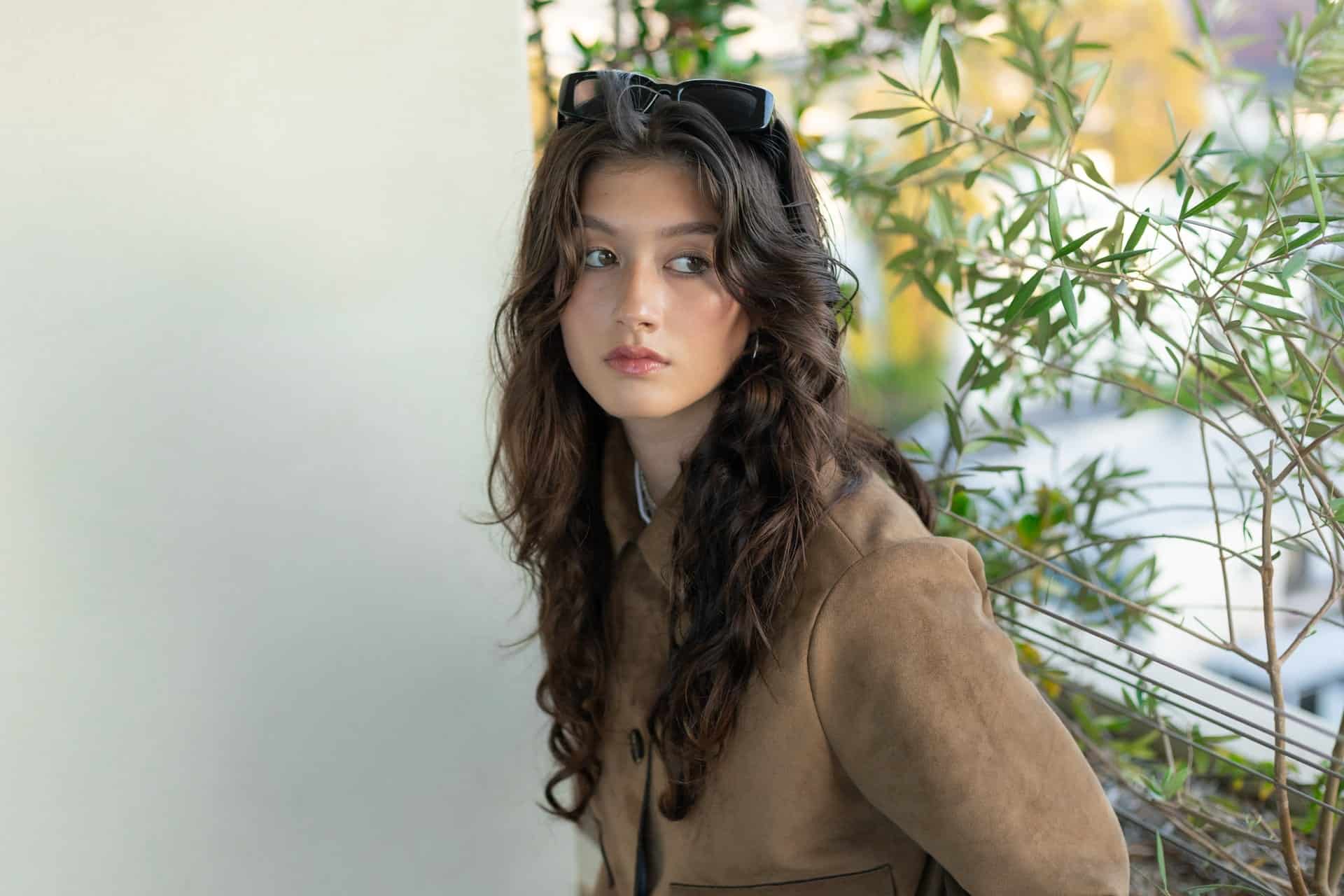 A young woman with long wavy brown hair, wearing a brown suede jacket and sunglasses on her head, looks to the side while standing near a wall and green leafy plants outdoors.