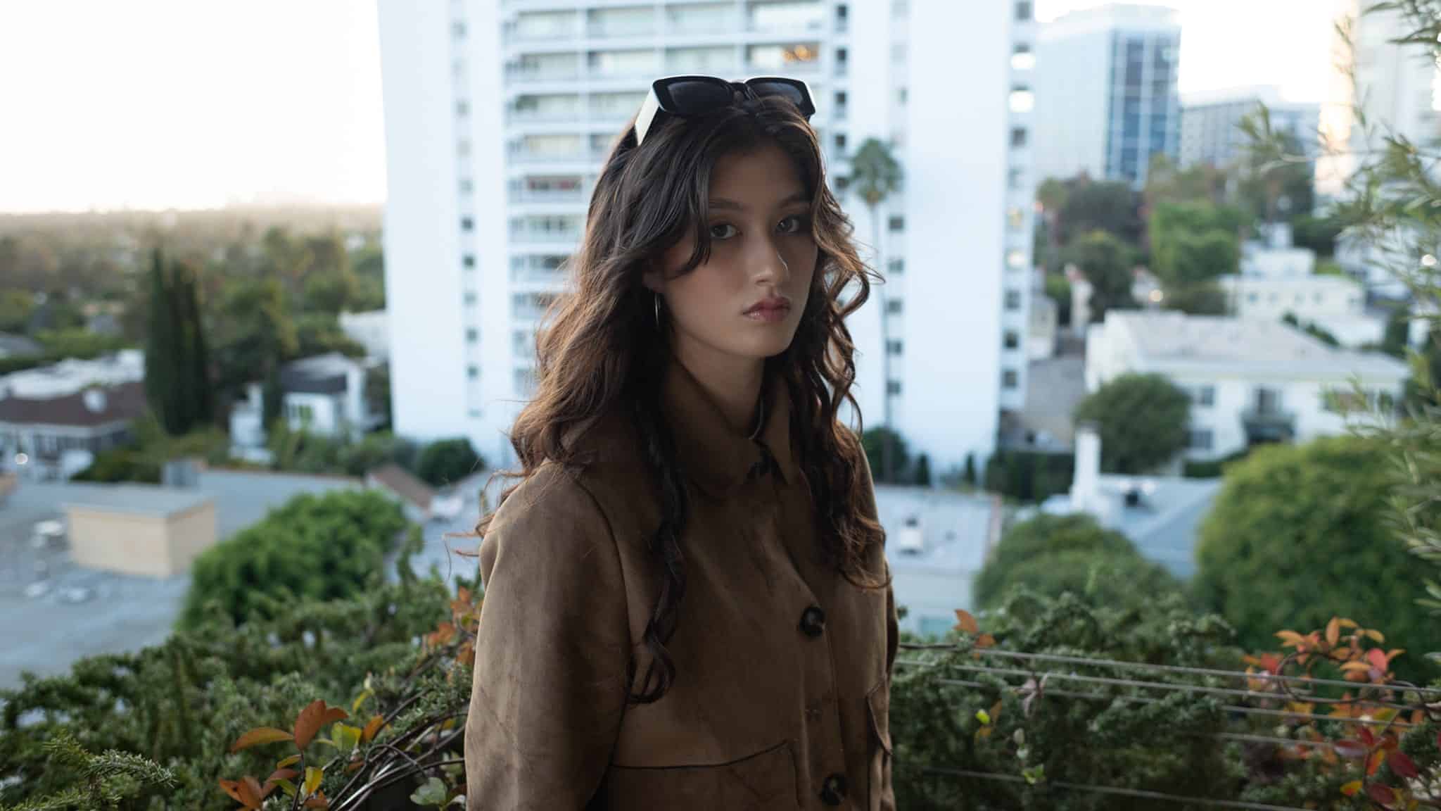 A young woman with long wavy brown hair, wearing a brown coat and sunglasses on her head, stands on a balcony with greenery. Tall white buildings and cityscape are visible in the background.