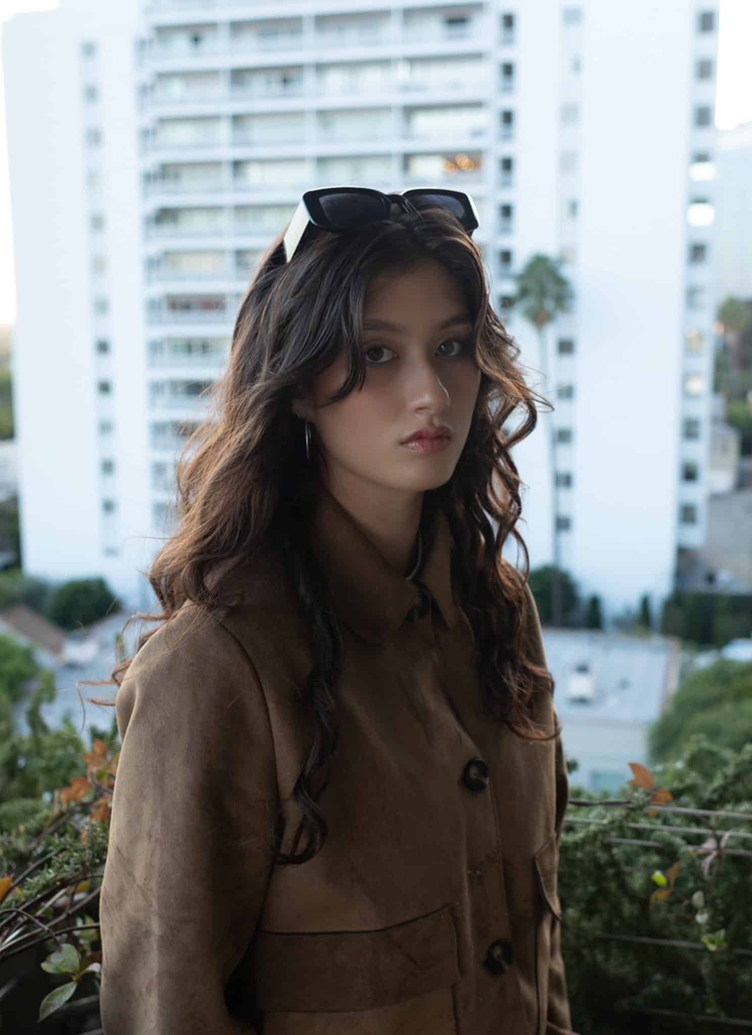 A young woman with long wavy brown hair, wearing a brown coat and sunglasses on her head, stands on a balcony with greenery. Tall white buildings and cityscape are visible in the background.