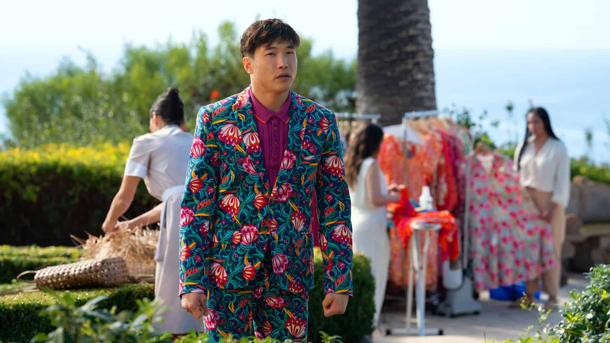 A man wearing a vibrant floral suit with a magenta shirt stands outdoors, looking ahead. Behind him, people arrange colorful clothes and dresses on racks near greenery and a palm tree, with the ocean visible in the background.