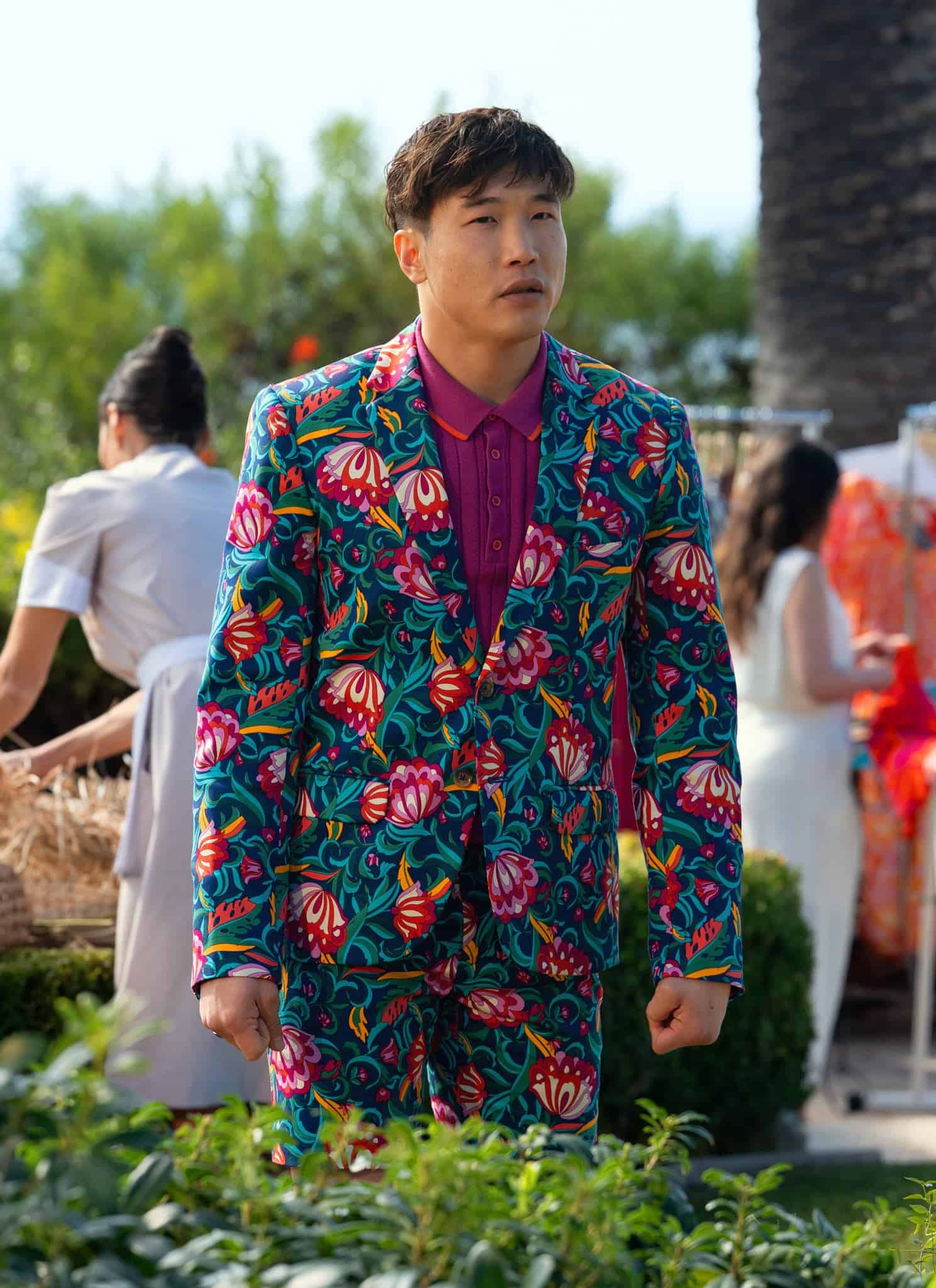A man wearing a vibrant floral suit with a magenta shirt stands outdoors, looking ahead. Behind him, people arrange colorful clothes and dresses on racks near greenery and a palm tree, with the ocean visible in the background.