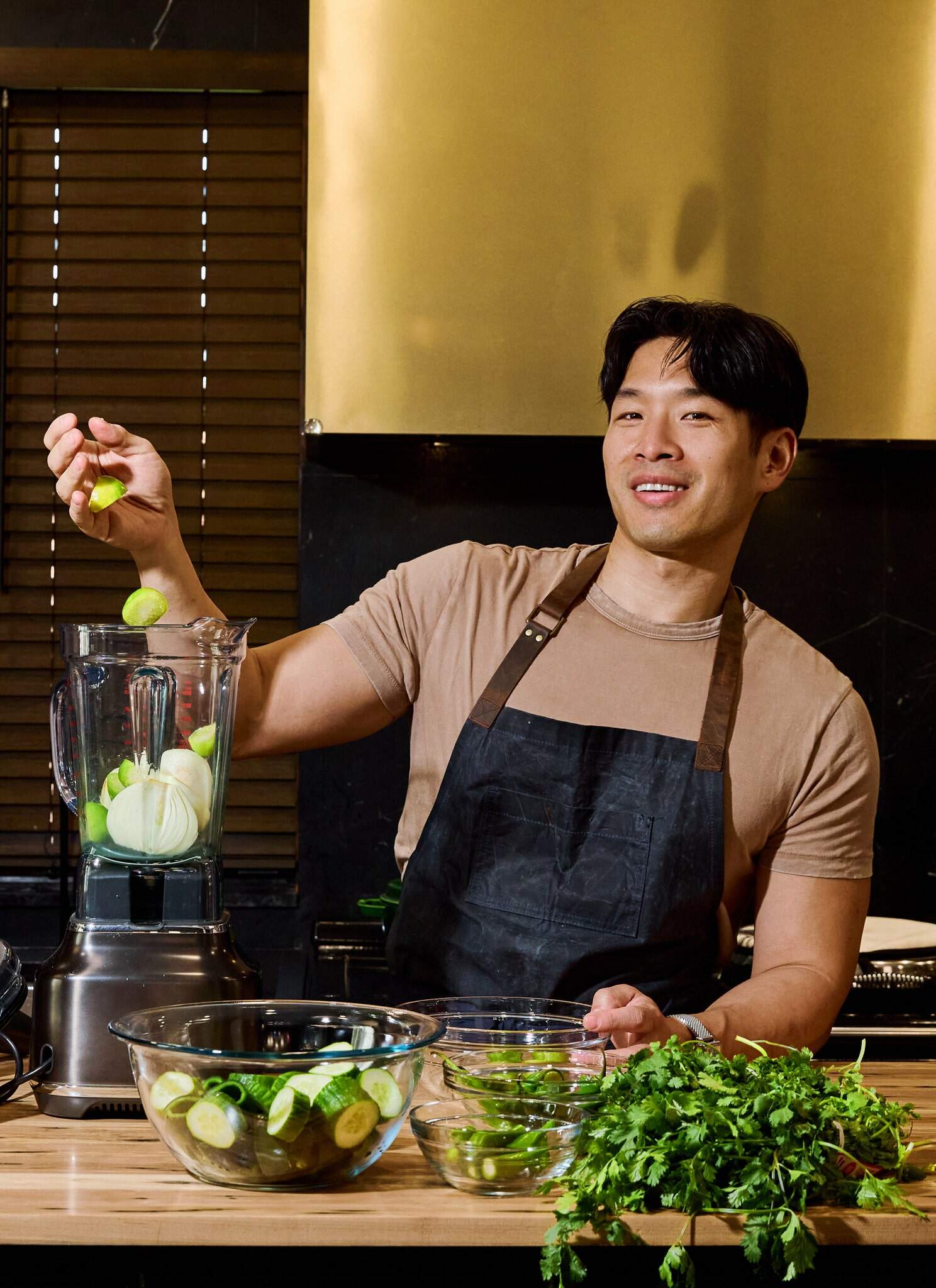 A person in a kitchen, wearing a brown shirt and a black apron, drops limes into a blender filled with other ingredients. Fresh herbs, cucumbers, and bowls are on the wooden countertop in front of them.