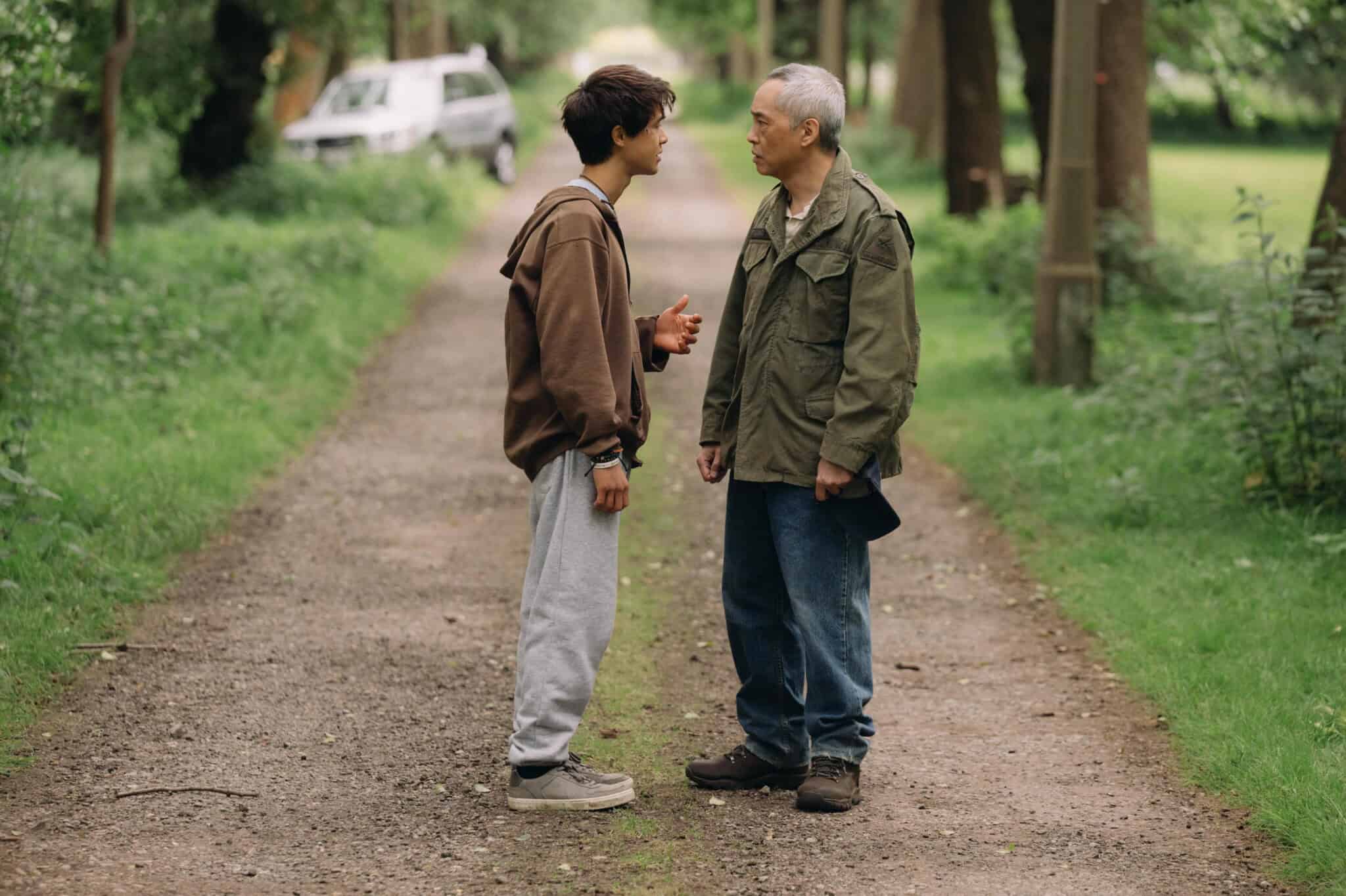 A young man and an older man stand facing each other on a tree-lined dirt path, engaged in conversation. A white car is parked in the background among the greenery.