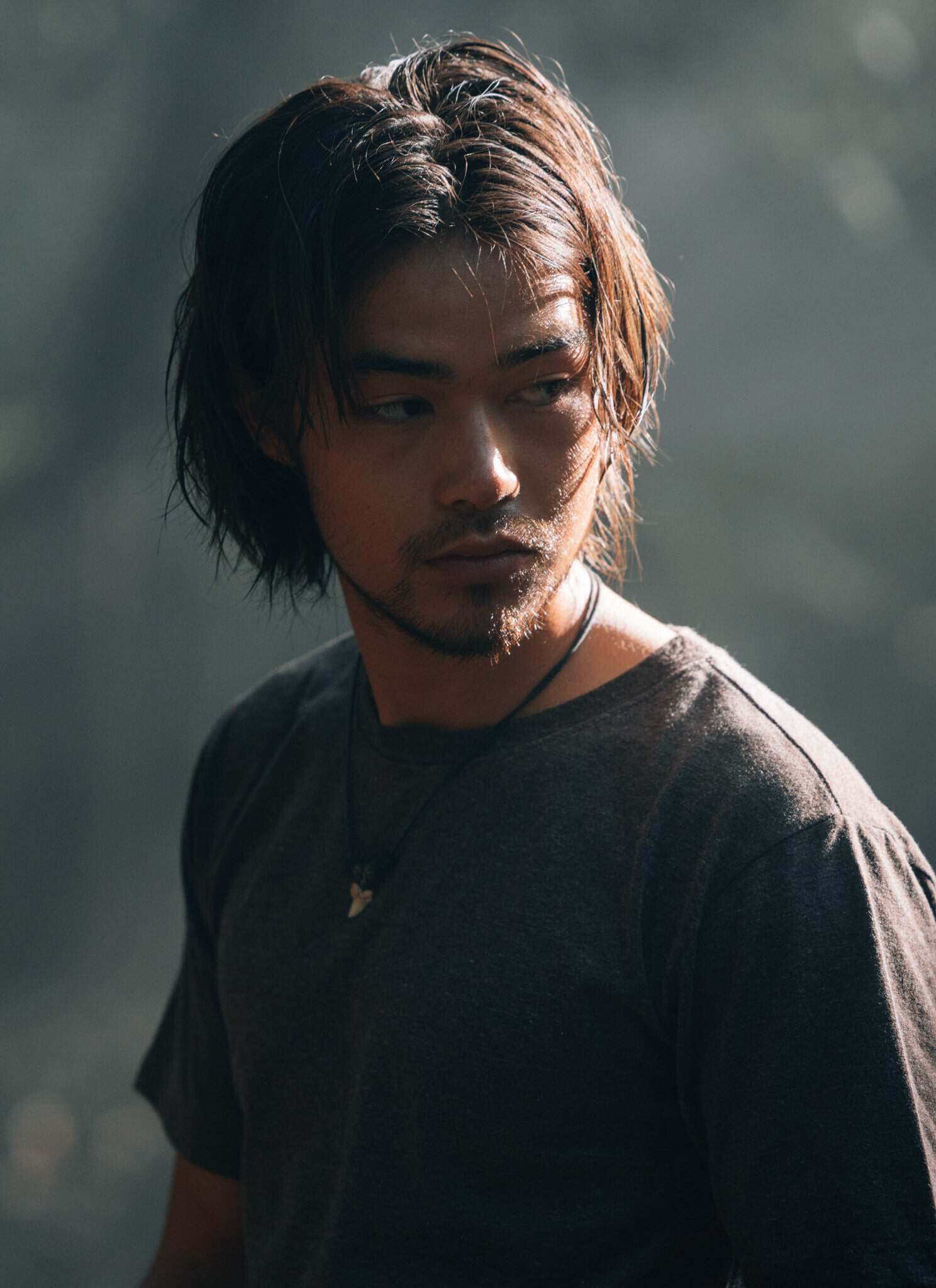 A young man with shoulder-length dark hair and a short beard stands in a forest, wearing a gray t-shirt and a necklace, looking to his right with a serious expression. Sunlight filters through the trees in the background.