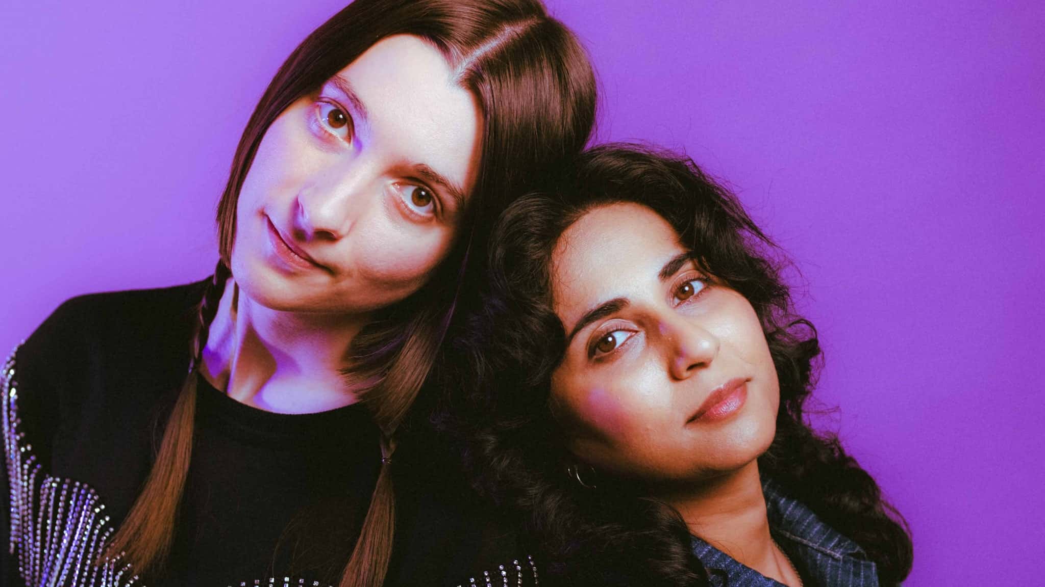 Two women pose closely together against a purple background; one has straight brown hair in pigtails and wears a black top, while the other has wavy dark hair and wears a denim shirt, both looking calmly at the camera.