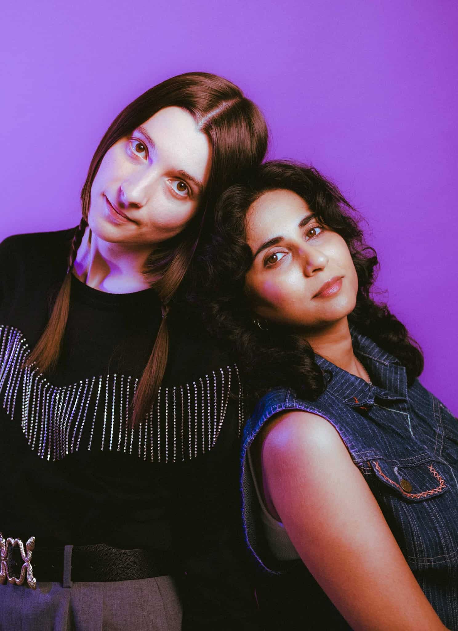 Two women pose closely together against a purple background; one has straight brown hair in pigtails and wears a black top, while the other has wavy dark hair and wears a denim shirt, both looking calmly at the camera.
