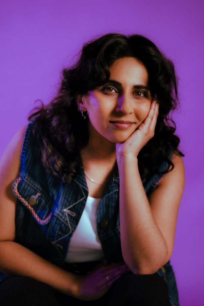 A person with wavy dark hair, wearing a denim vest over a white top, rests their face on their hand and smiles slightly, sitting against a purple background.