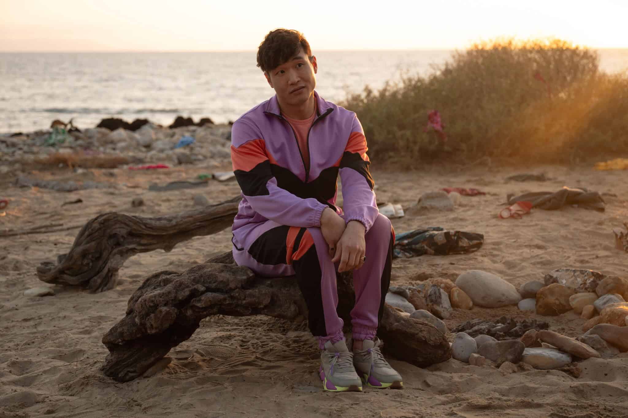 A person in a purple and pink tracksuit sits on a driftwood log at a sandy beach during sunset, with the ocean and scattered rocks and vegetation in the background.