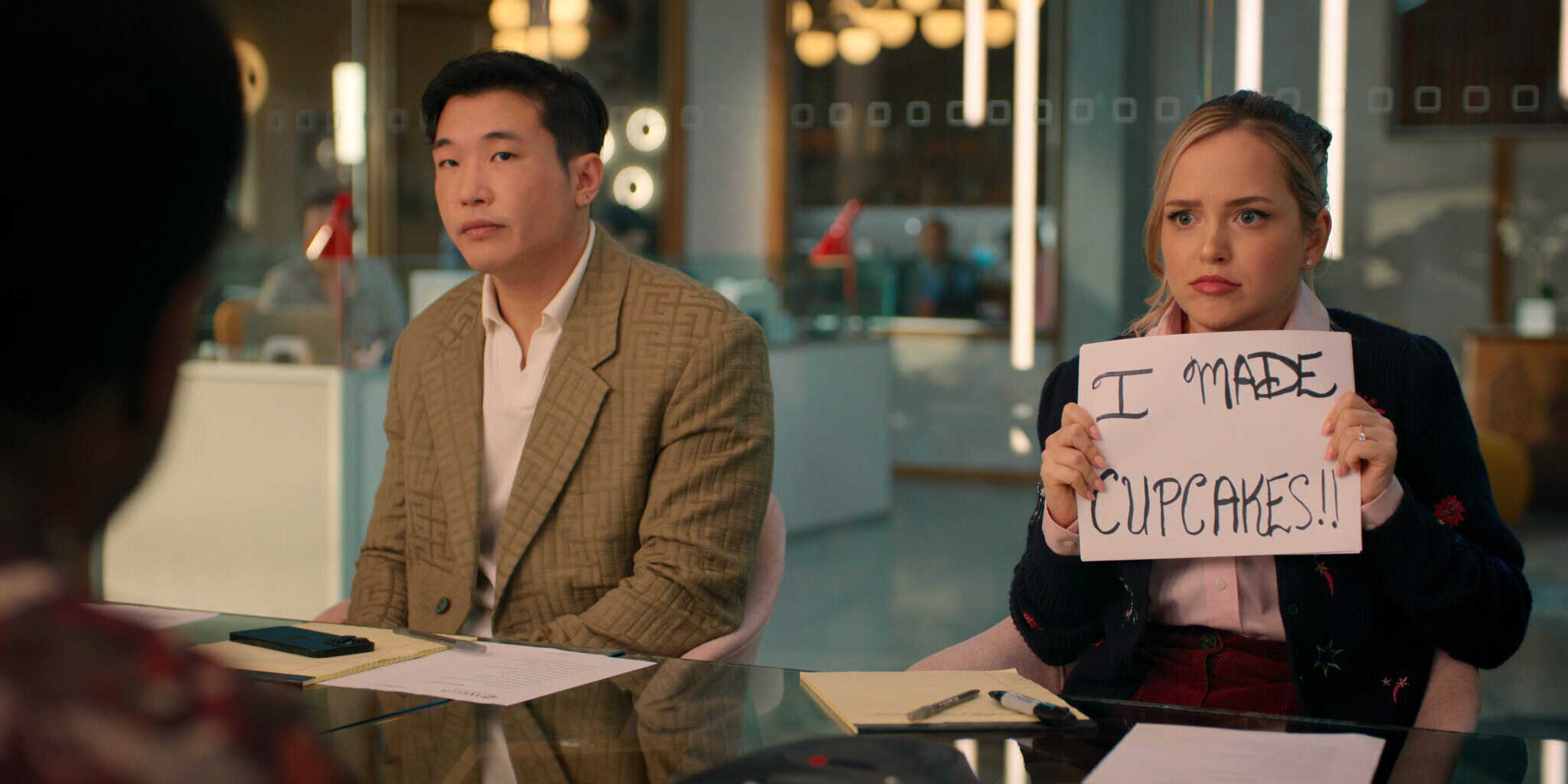 A woman sitting at a conference table holds up a handwritten sign that says I MADE CUPCAKES!! while a man sits beside her, both looking serious. Office setting with papers and laptops on the table.