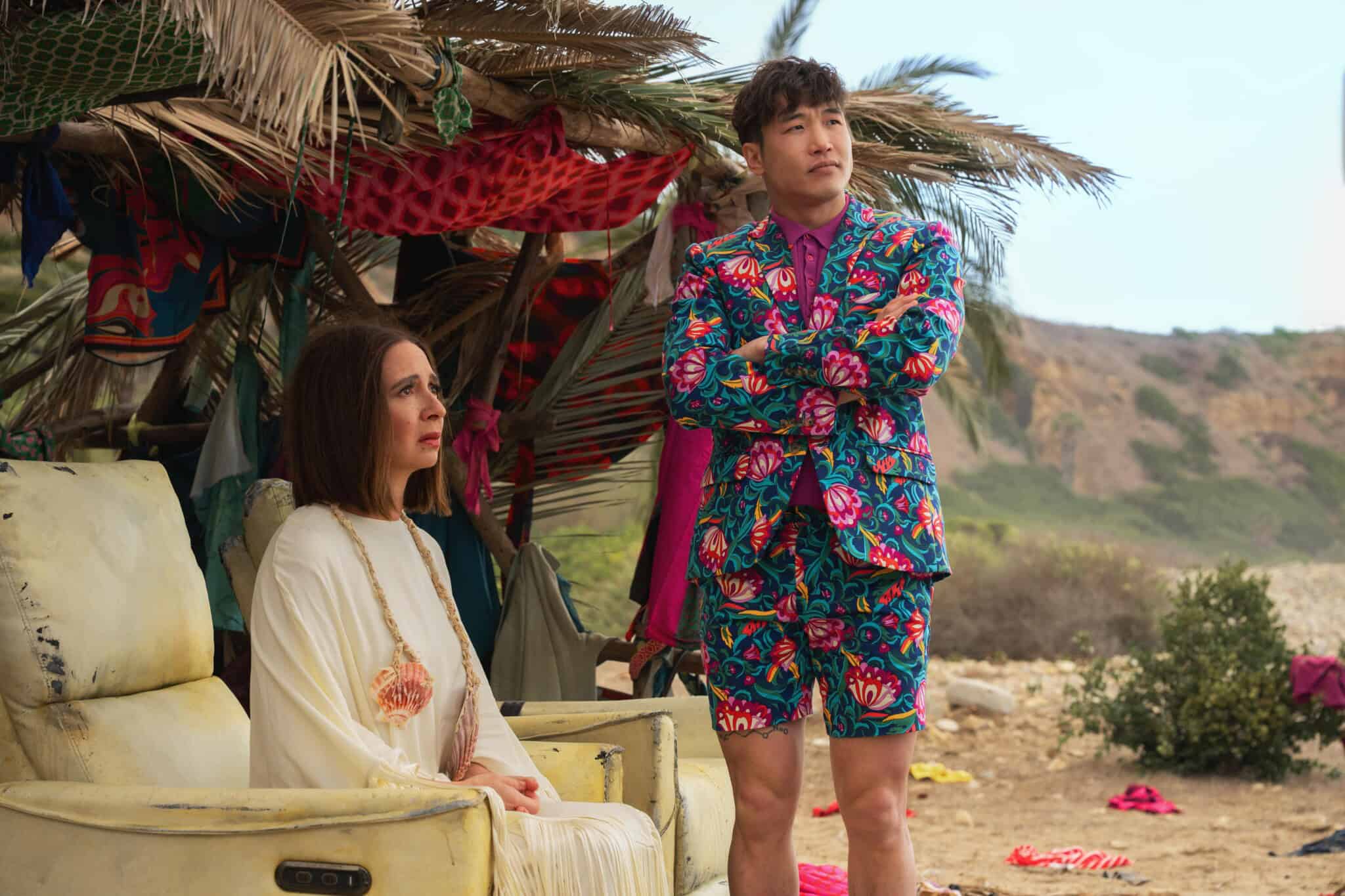 A woman in a white dress sits on a worn armchair beside a man standing with crossed arms, wearing a brightly colored floral suit and shorts. They are under a palm-covered shelter on a rocky beach.