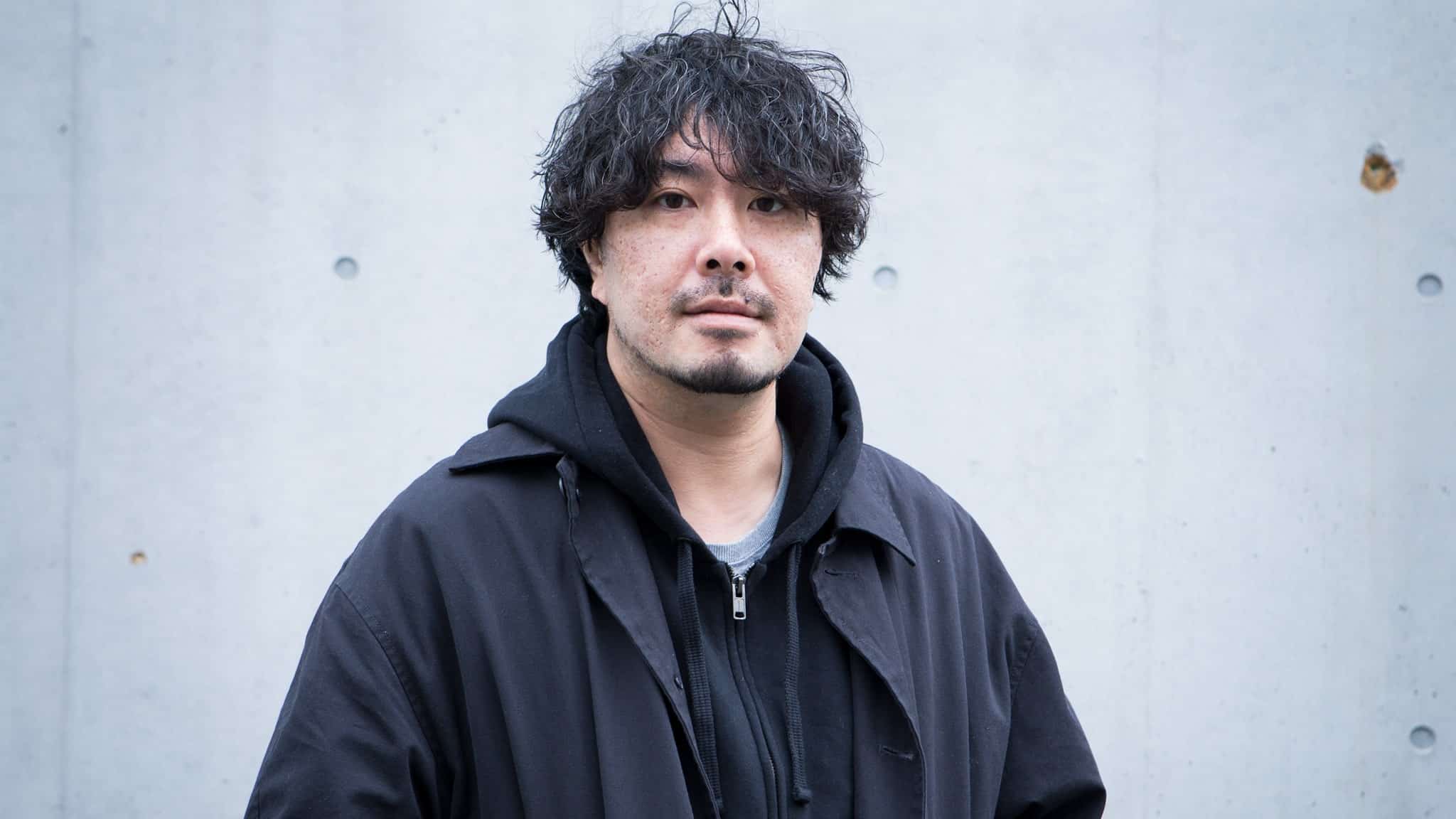 A man with wavy dark hair and facial hair stands in front of a light gray concrete wall, wearing a black coat over a black hoodie. He has a neutral expression and looks directly at the camera.