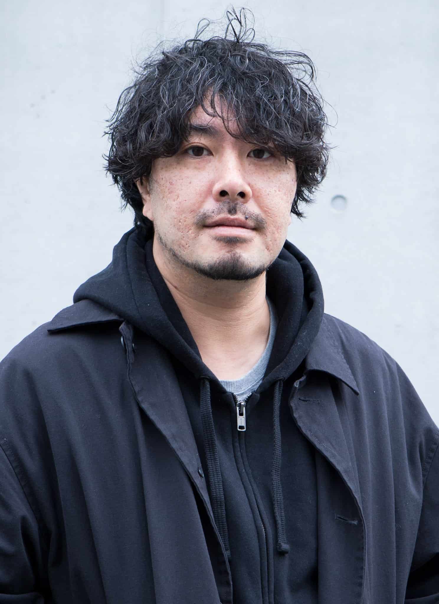 A man with wavy dark hair and facial hair stands in front of a light gray concrete wall, wearing a black coat over a black hoodie. He has a neutral expression and looks directly at the camera.
