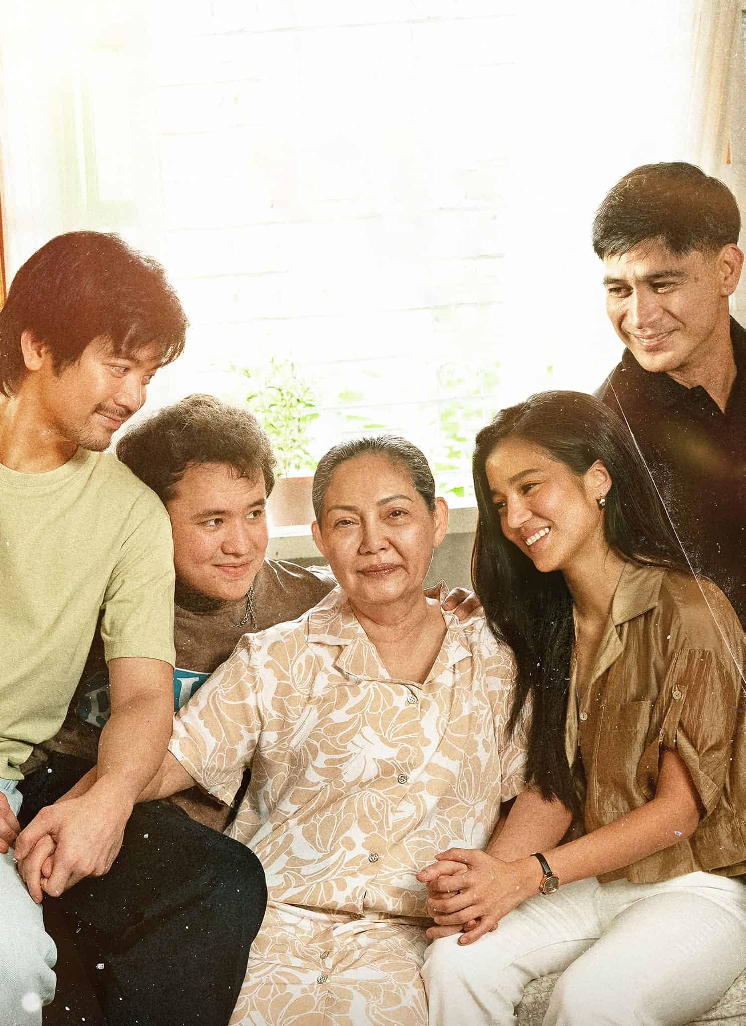 Five people of different ages sit closely together and smile warmly in a sunlit room. An older woman is in the center, surrounded by two men and two younger adults, creating a sense of family and togetherness.