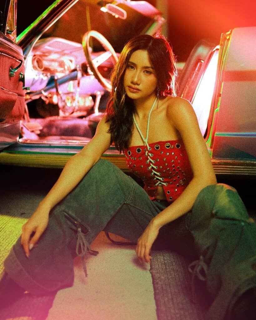 A woman with long dark hair sits on the ground by an open car door, wearing a red lace-up bandeau top and loose-fitting cargo pants. Neon lights cast a colorful glow on the scene, creating a vibrant, retro atmosphere.