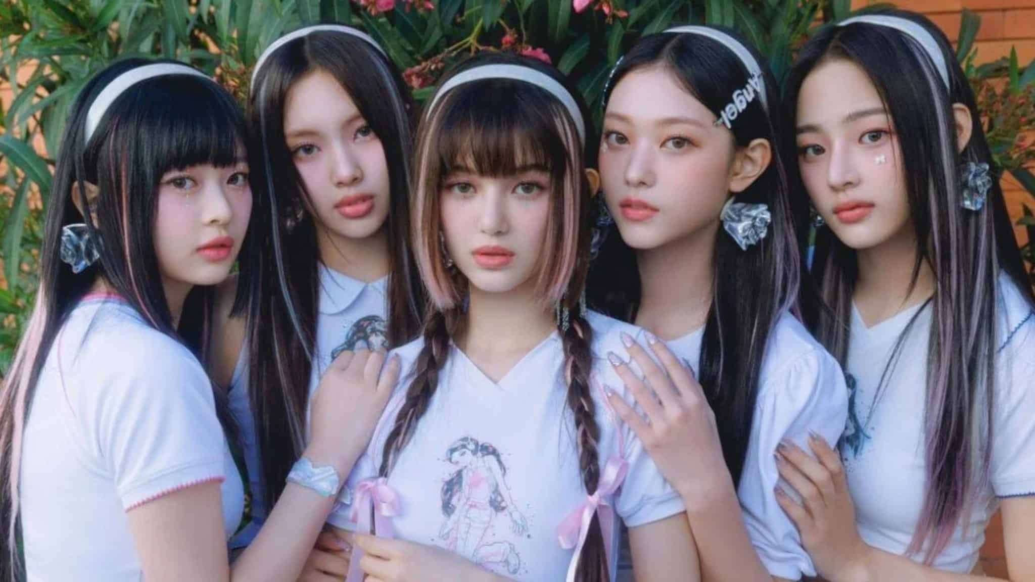 Five young women with long, straight hair and matching white shirts pose closely together outdoors, surrounded by greenery. They wear headbands and have neutral expressions, looking directly at the camera.