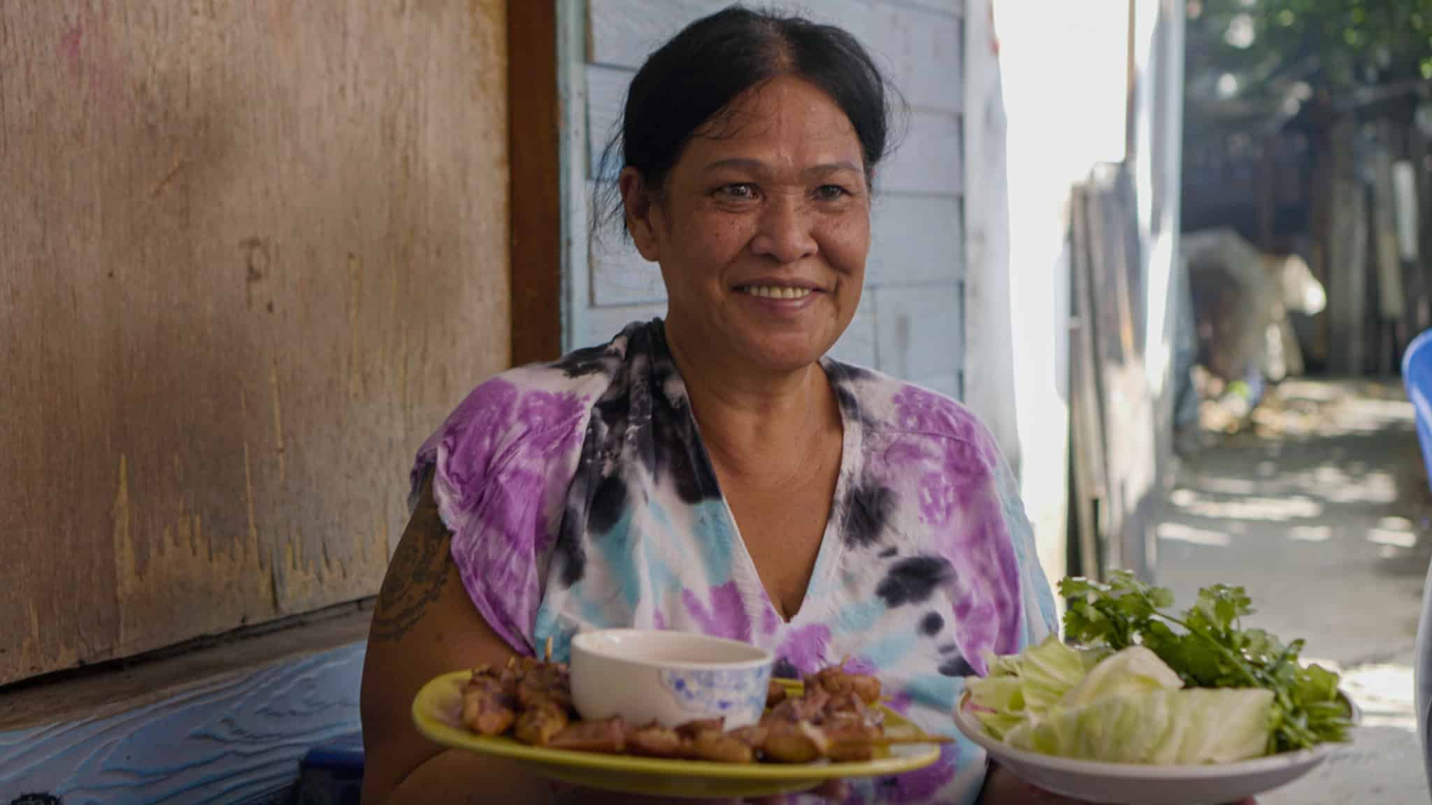 A smiling woman with dark hair in a tie-dye shirt holds two plates: one with grilled meat and a dipping sauce, the other with fresh vegetables. She sits outside near a wooden wall with sunlight in the background.