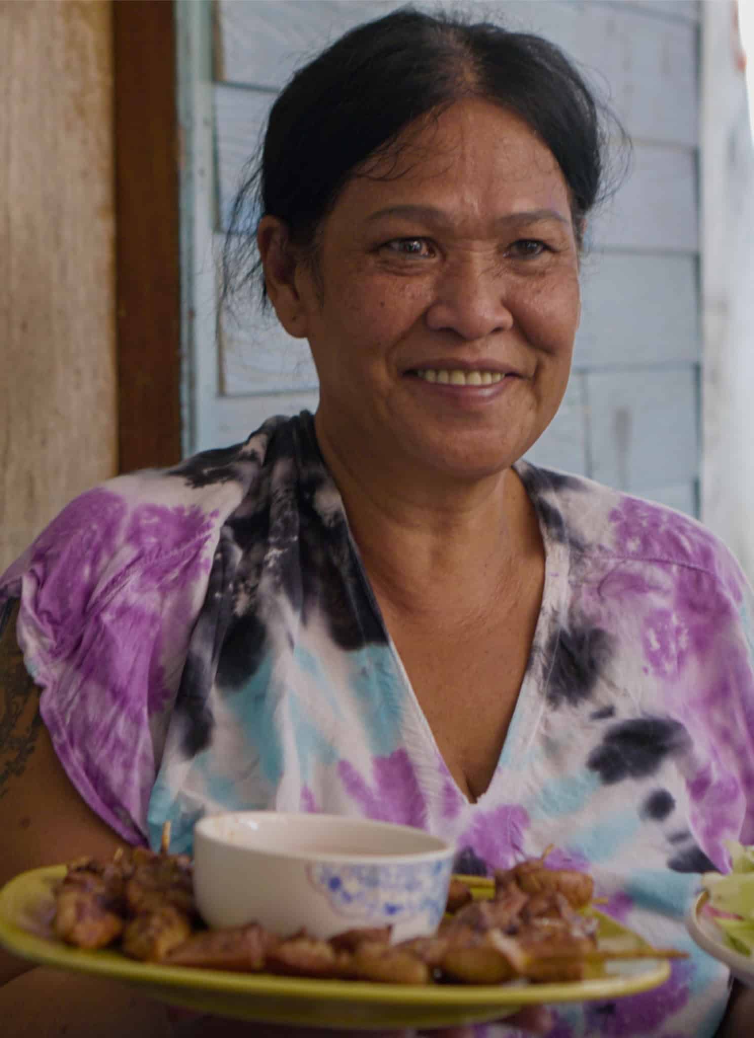 A smiling woman with dark hair in a tie-dye shirt holds two plates: one with grilled meat and a dipping sauce, the other with fresh vegetables. She sits outside near a wooden wall with sunlight in the background.