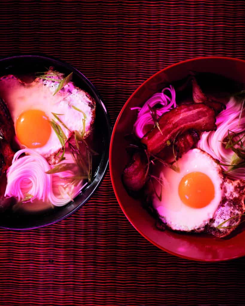 Two bowls of noodle soup topped with sunny-side-up eggs, crispy bacon, pink pickled onions, and green onions, placed on a striped textured surface with warm, vibrant lighting.