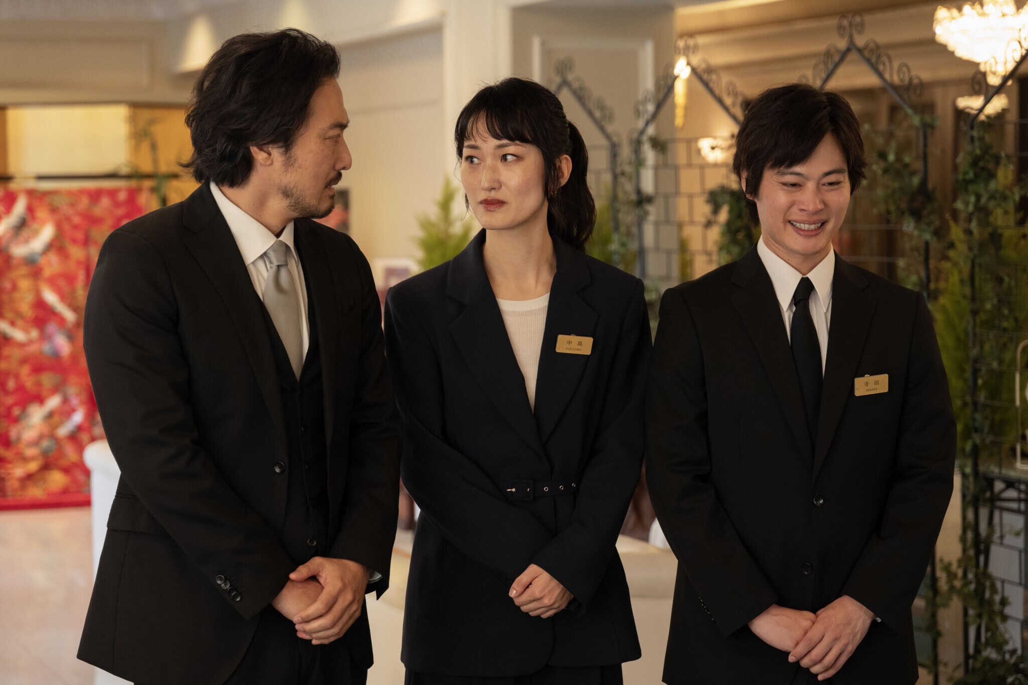 Three people in suits with name tags stand indoors; the man on the left speaks to the woman in the center while the man on the right smiles. The setting appears formal, with plants and decorations in the background.