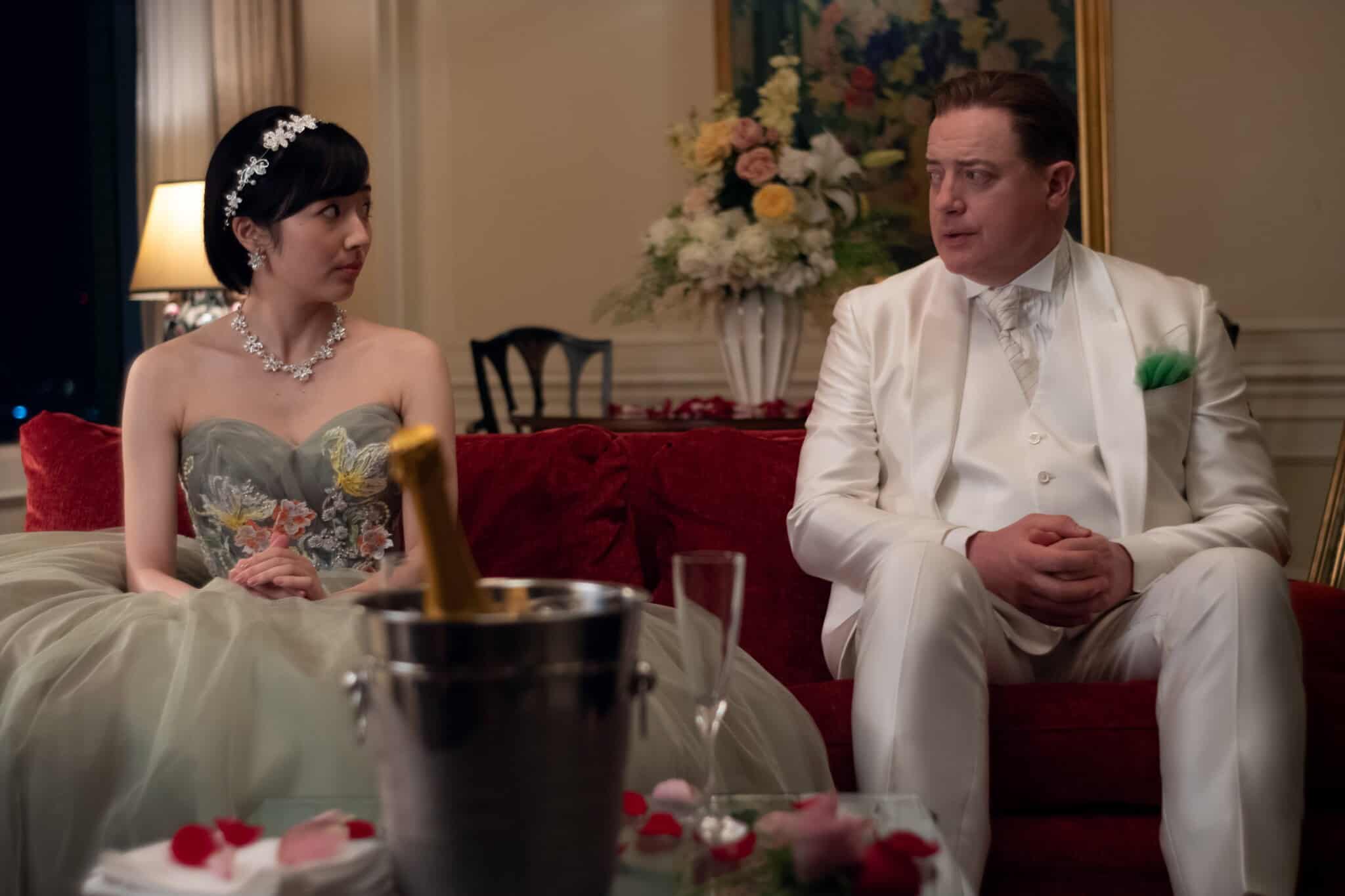 A woman in a floral strapless gown and a man in a white suit sit on a red couch, looking at each other. A champagne bottle, glass, and rose petals are on the table in front of them in an elegantly decorated room.