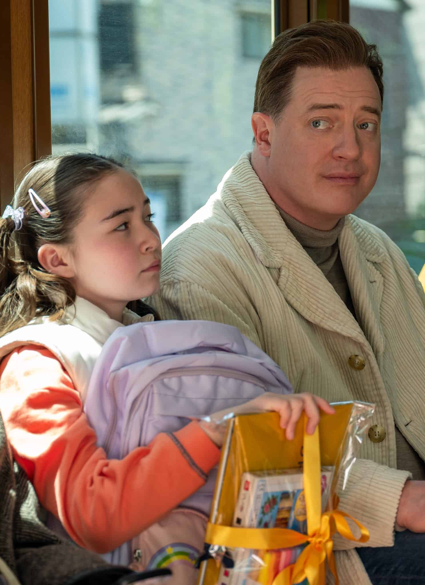 A man in a cream jacket sits next to a young girl with a purple backpack and orange sweater on a bus. They are surrounded by other passengers, all seated and looking in different directions.