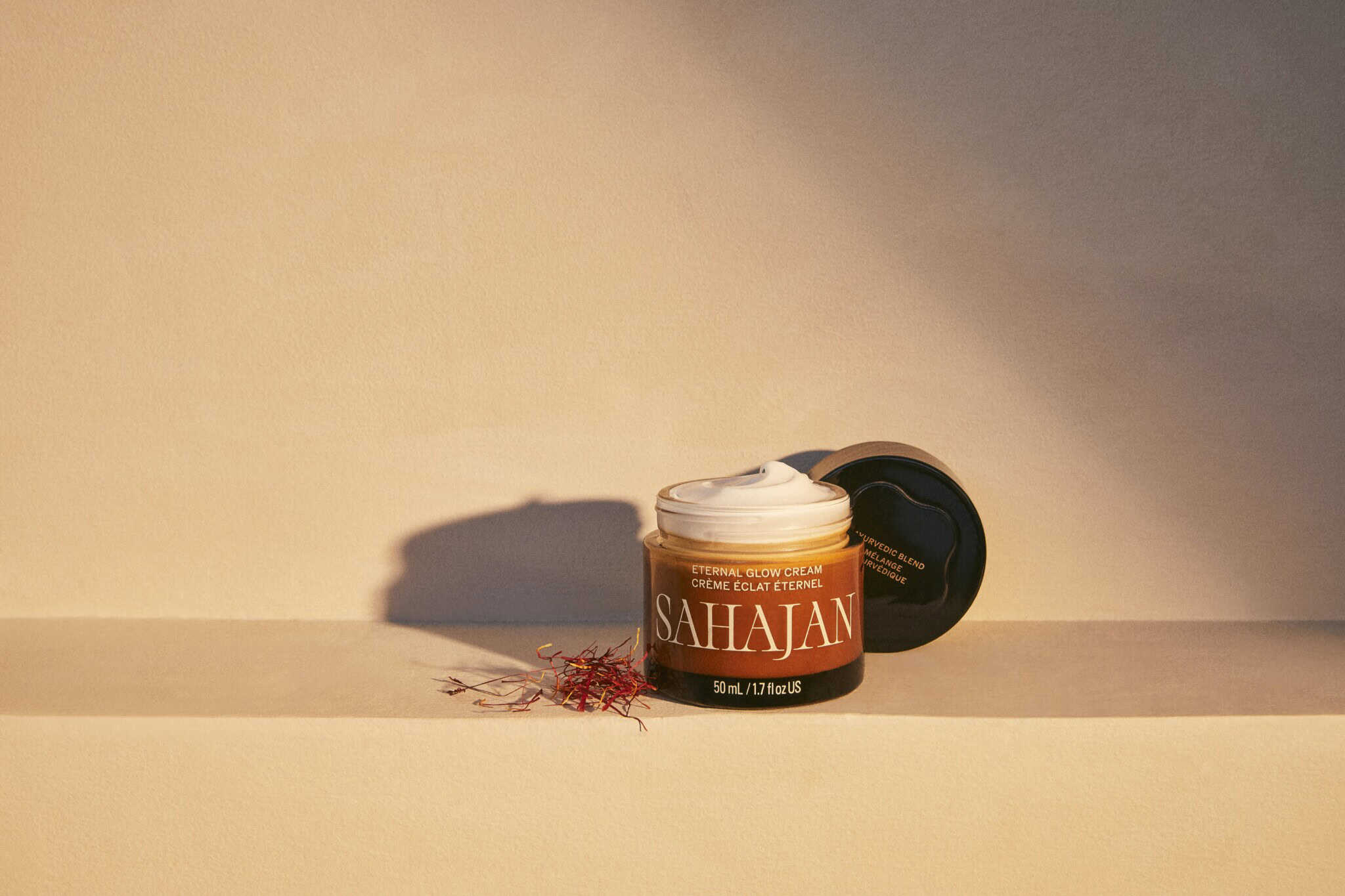 A jar of Sahajan Radiance Eye Cream with its black lid resting beside it on a beige surface, some cream visible at the top, and saffron strands scattered nearby, all lit by warm, soft light.