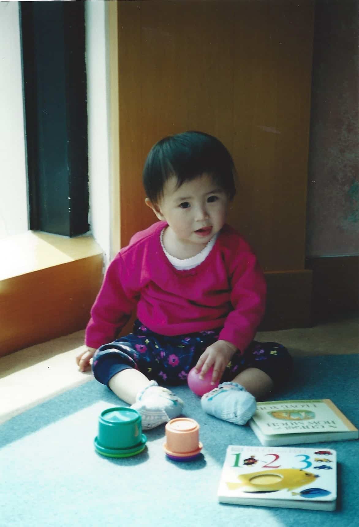 A young child in a pink sweater sits on the floor by a window, playing with stacking cups and a counting book. The child has short dark hair and appears focused and curious.