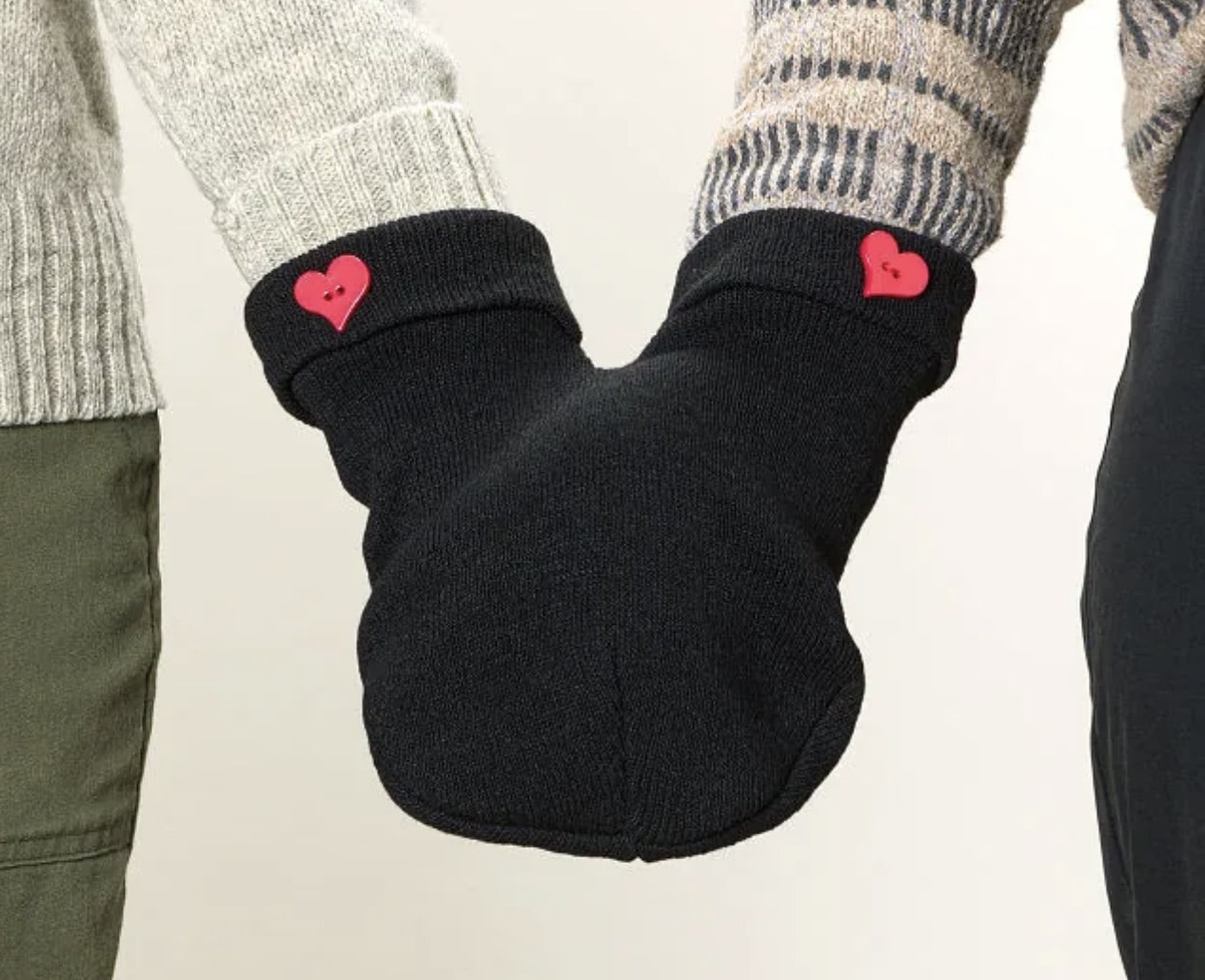 Two people wear a single pair of connected black mittens with red heart buttons, holding hands. Their sweaters and pants are visible from the elbows down against a neutral background.