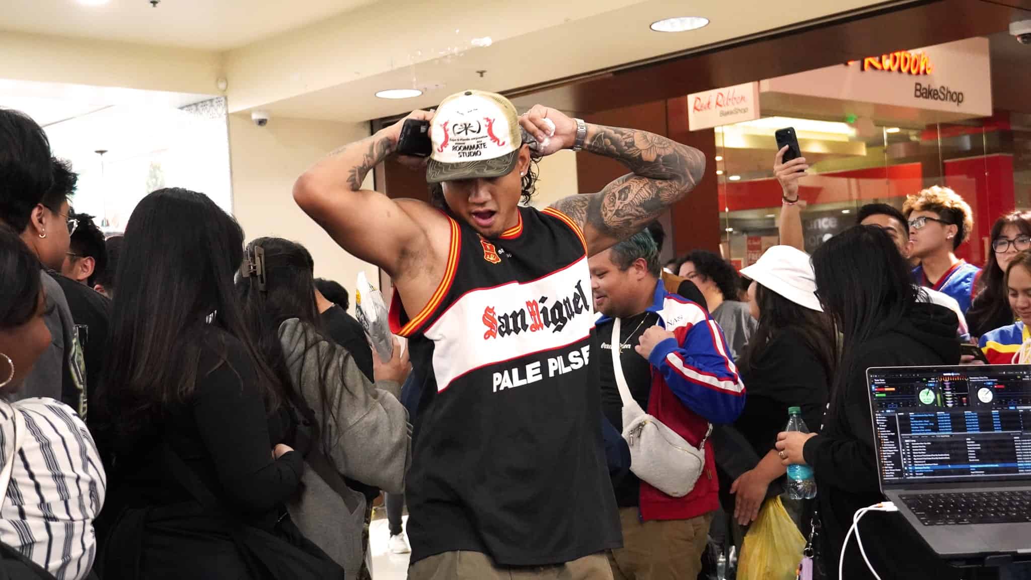 A man in a black tank top and cap dances energetically in a crowded indoor space. People surround him, some filming or watching, and a DJ setup is visible in the foreground. The atmosphere is lively and festive.