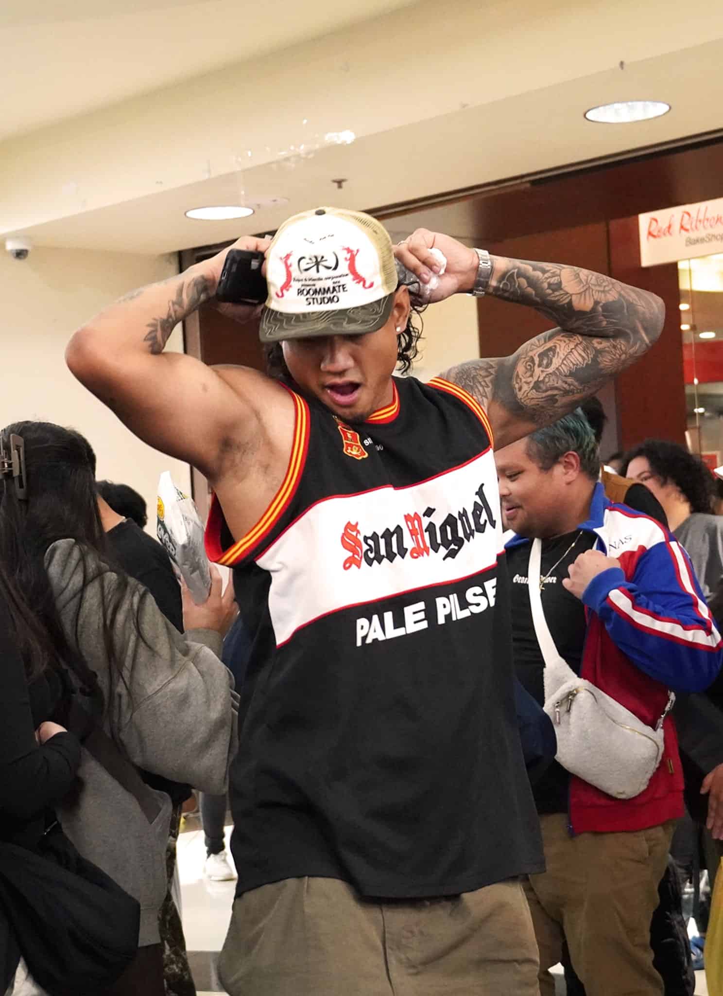 A man in a black tank top and cap dances energetically in a crowded indoor space. People surround him, some filming or watching, and a DJ setup is visible in the foreground. The atmosphere is lively and festive.