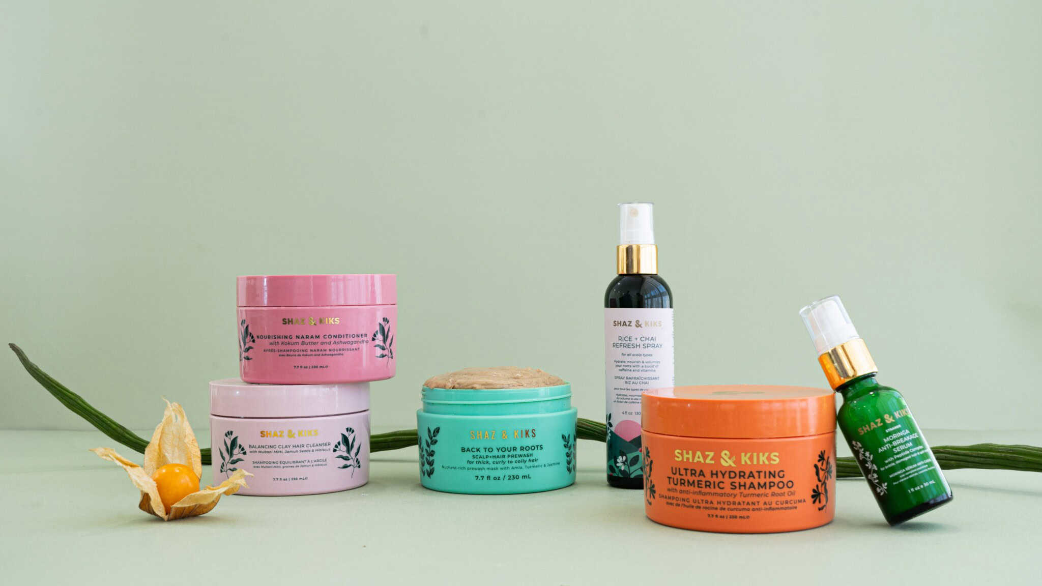 Six colorful hair care products in jars and bottles are arranged on a light green surface with aloe vera leaves and a yellow fruit, against a pale green background. The products have pink, orange, green, and blue packaging.