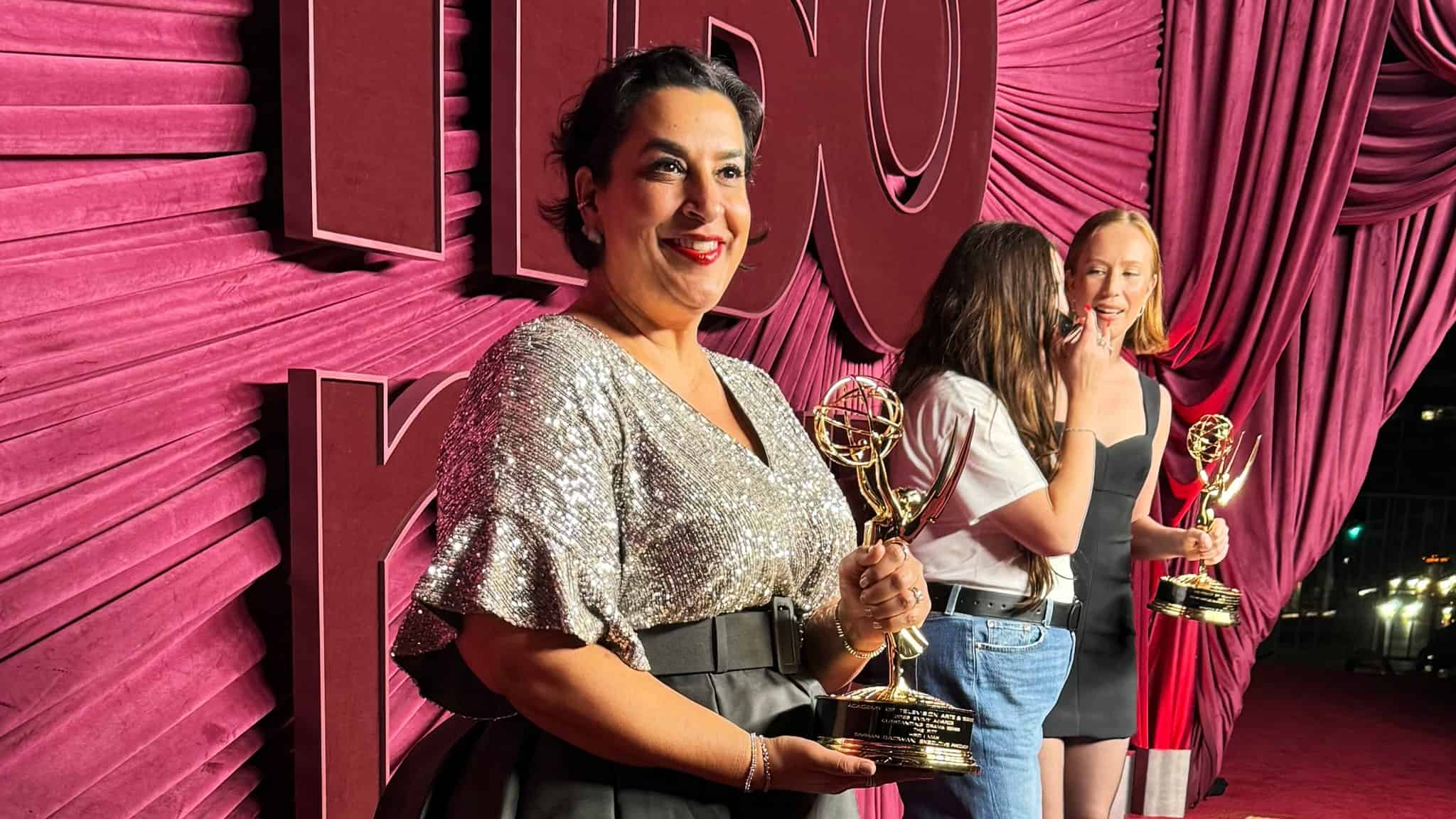 Three women stand on a red carpet with a draped red background, each holding an Emmy award. The woman in front wears a sparkly silver top and black skirt, smiling at the camera. The other two are in conversation in the background.