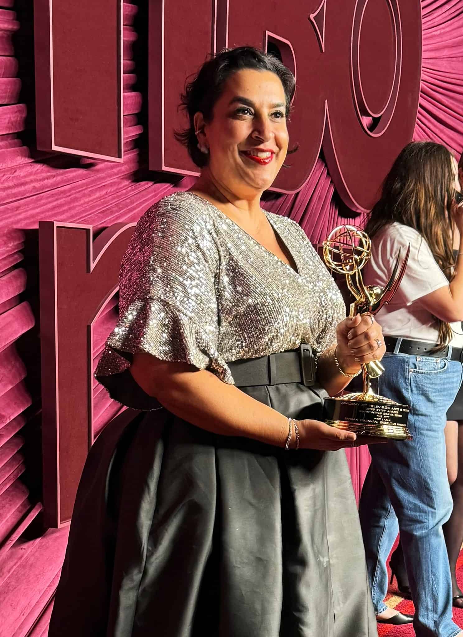 Three women stand on a red carpet with a draped red background, each holding an Emmy award. The woman in front wears a sparkly silver top and black skirt, smiling at the camera. The other two are in conversation in the background.
