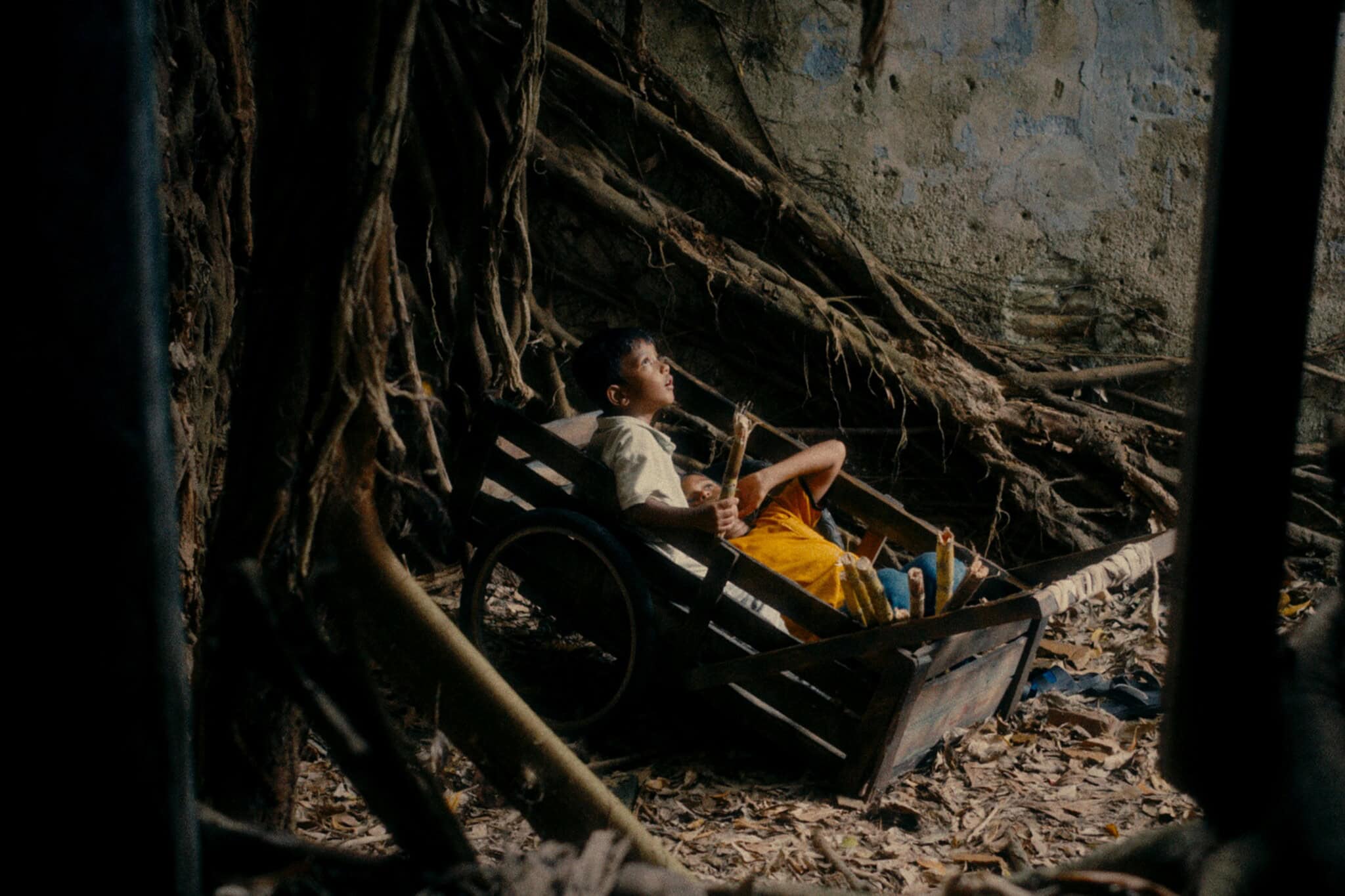 A young boy in a yellow shirt and orange shorts lies in a wooden cart, gazing upward among large tree roots and fallen leaves in a dilapidated, overgrown area.