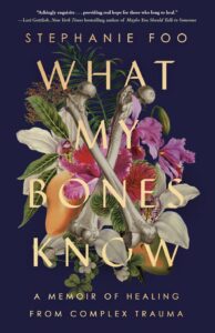 Book cover of What My Bones Know by Stephanie Foo. The title is overlaid on a bouquet of colorful flowers and two crossed bones, all set against a dark background. Subheading reads: A Memoir of Healing from Complex Trauma.