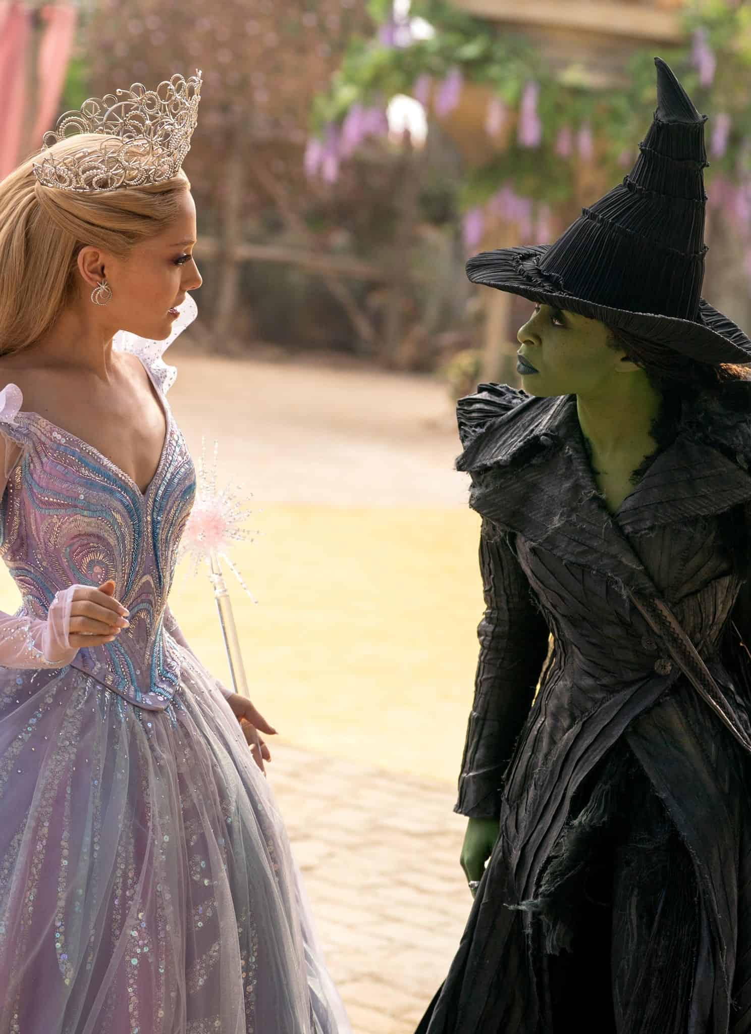 A woman in a sparkling, pastel-colored ball gown and crown faces another woman dressed as a green-skinned witch in black robes and a pointed hat, holding a staff, standing on a yellow brick road in a magical setting.